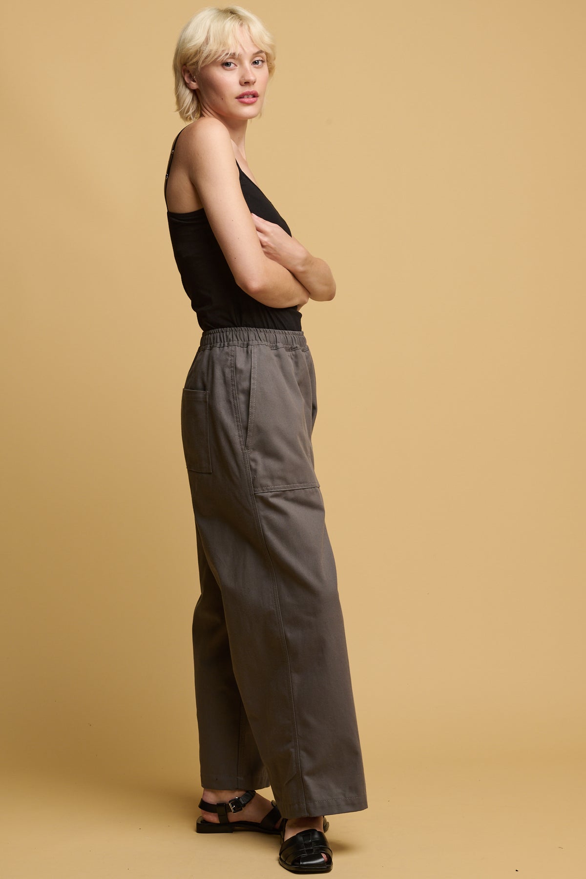 The side of female wearing camerawoman pants in charcoal paired with black camisole tucked in and black sandals