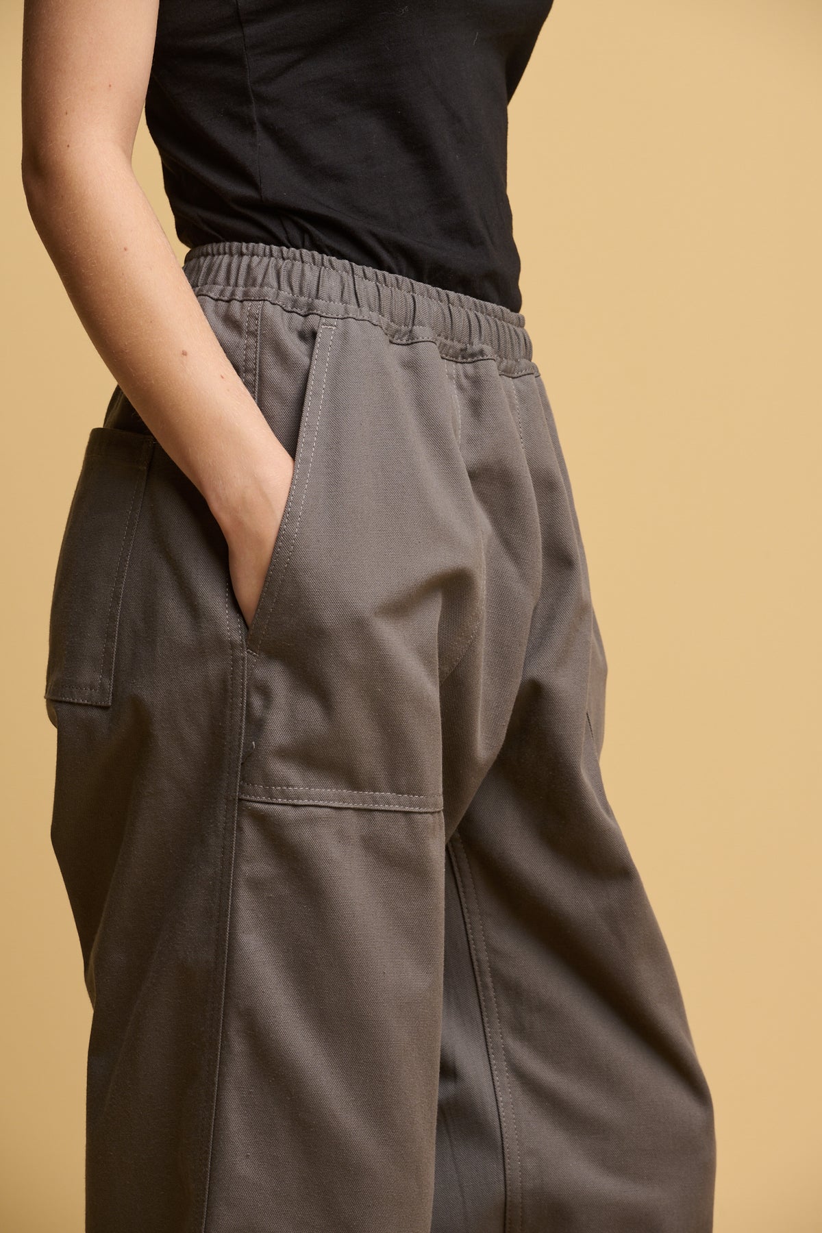 Close up of female model with her hands in the pocket of camerawoman drawstring tapered cotton canvas trousers in charcoal with a black camisole tucked in