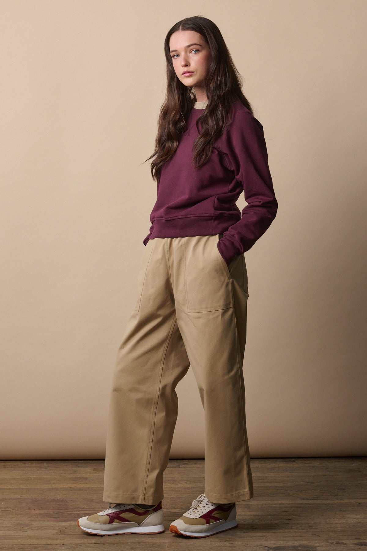 
            Full body shot of brunette female wearing camerawoman trousers in putty from the side showing the wide tapered shape of the leg. Worn with raglan sweatshirt in plum