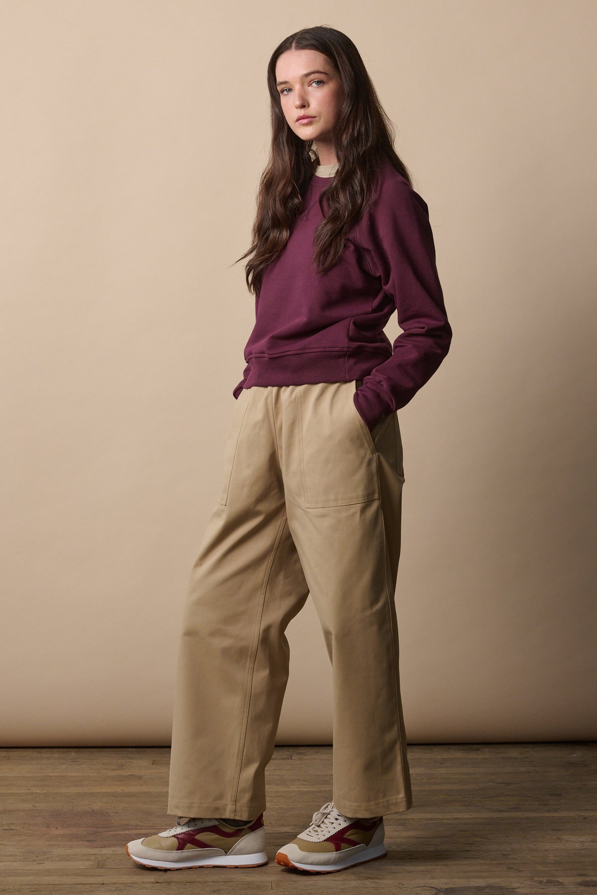 Full body shot of brunette female wearing camerawoman trousers in putty from the side showing the wide tapered shape of the leg. Worn with raglan sweatshirt in plum