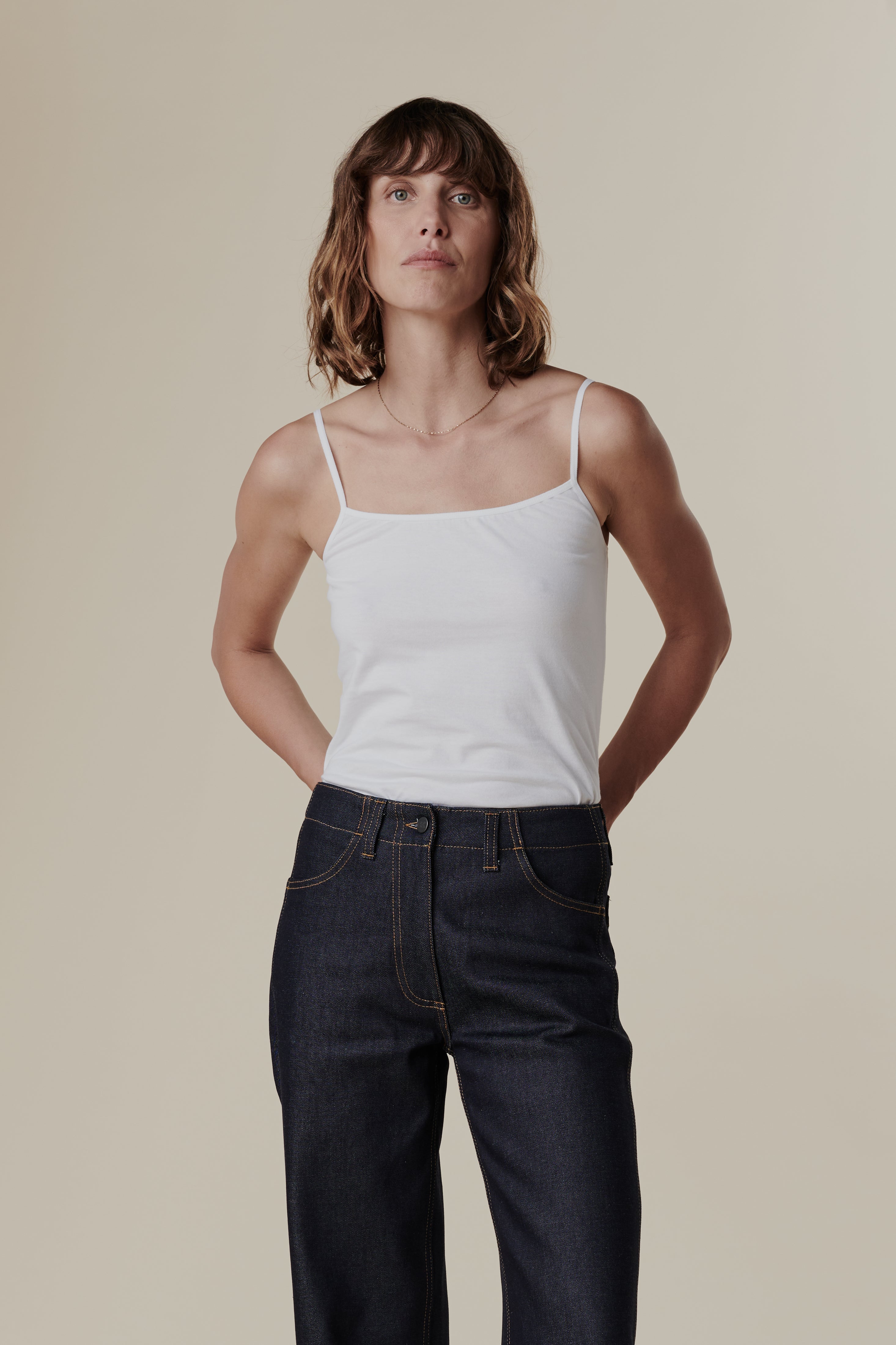 Thigh up front of female with shoulder length hair wearing white camisole tucked into work jeans in indigo 