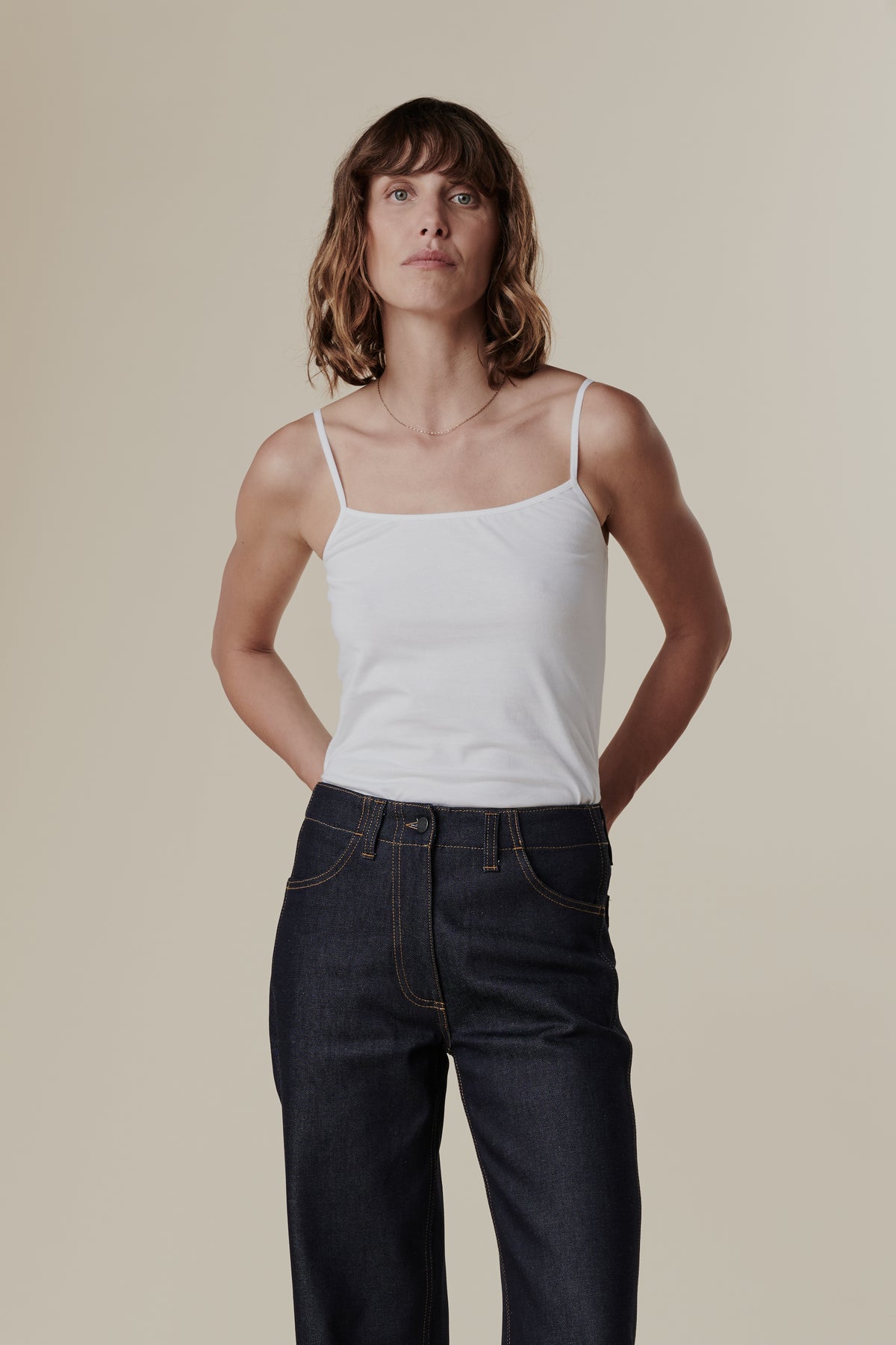 Thigh up front of female with shoulder length hair wearing white camisole tucked into work jeans in indigo
