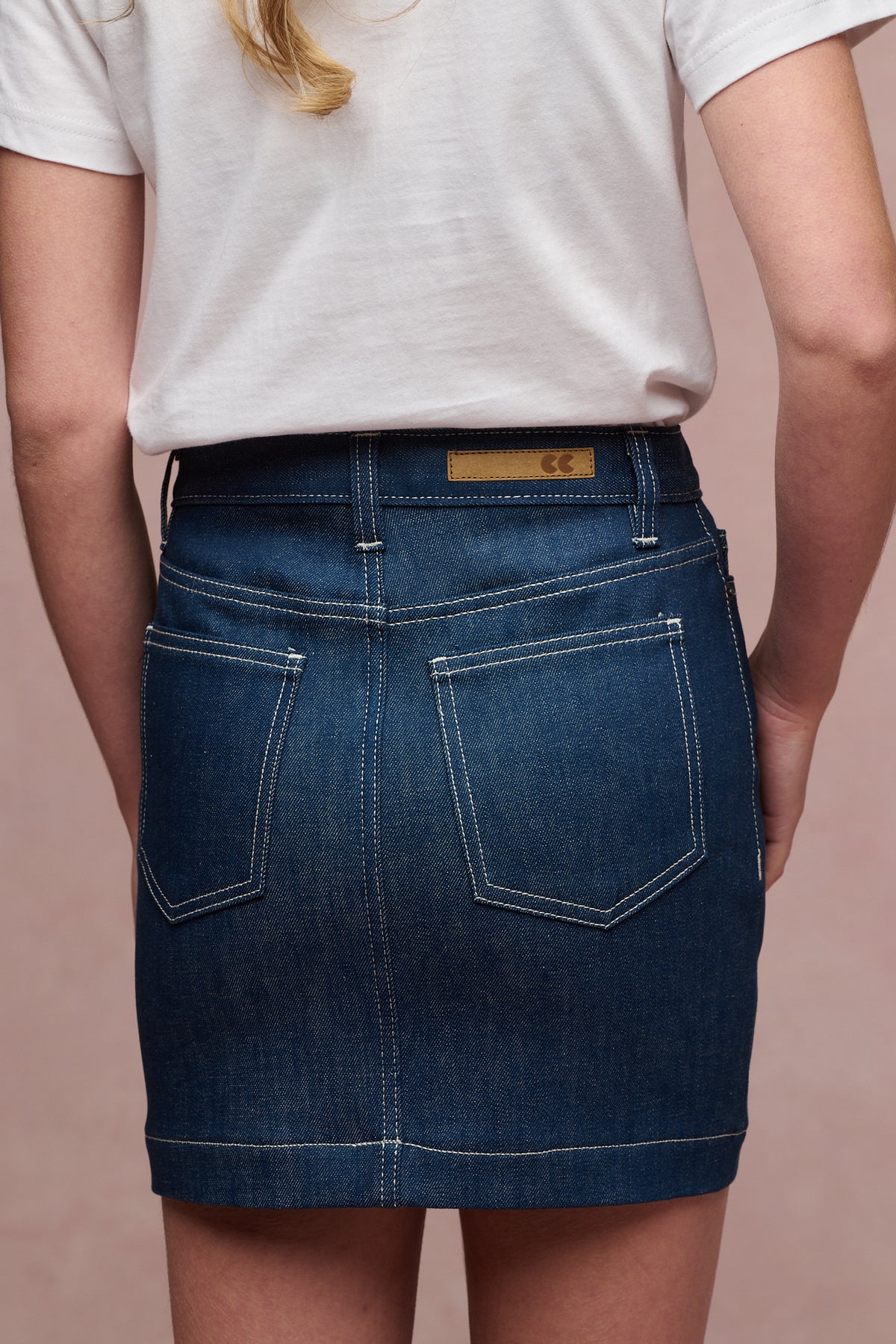 Close up of the back of female in Charlie denim mini skirt. Two pack pockets and belt loops, CC logo jean patch on waistband