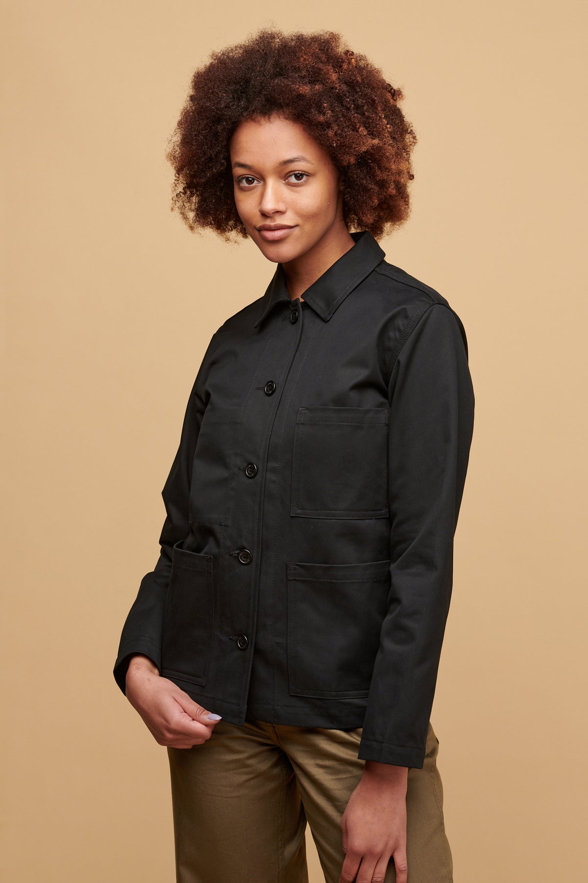 Thigh up of female wearing chore jacket in black with the buttons fastened, three external front pockets.
