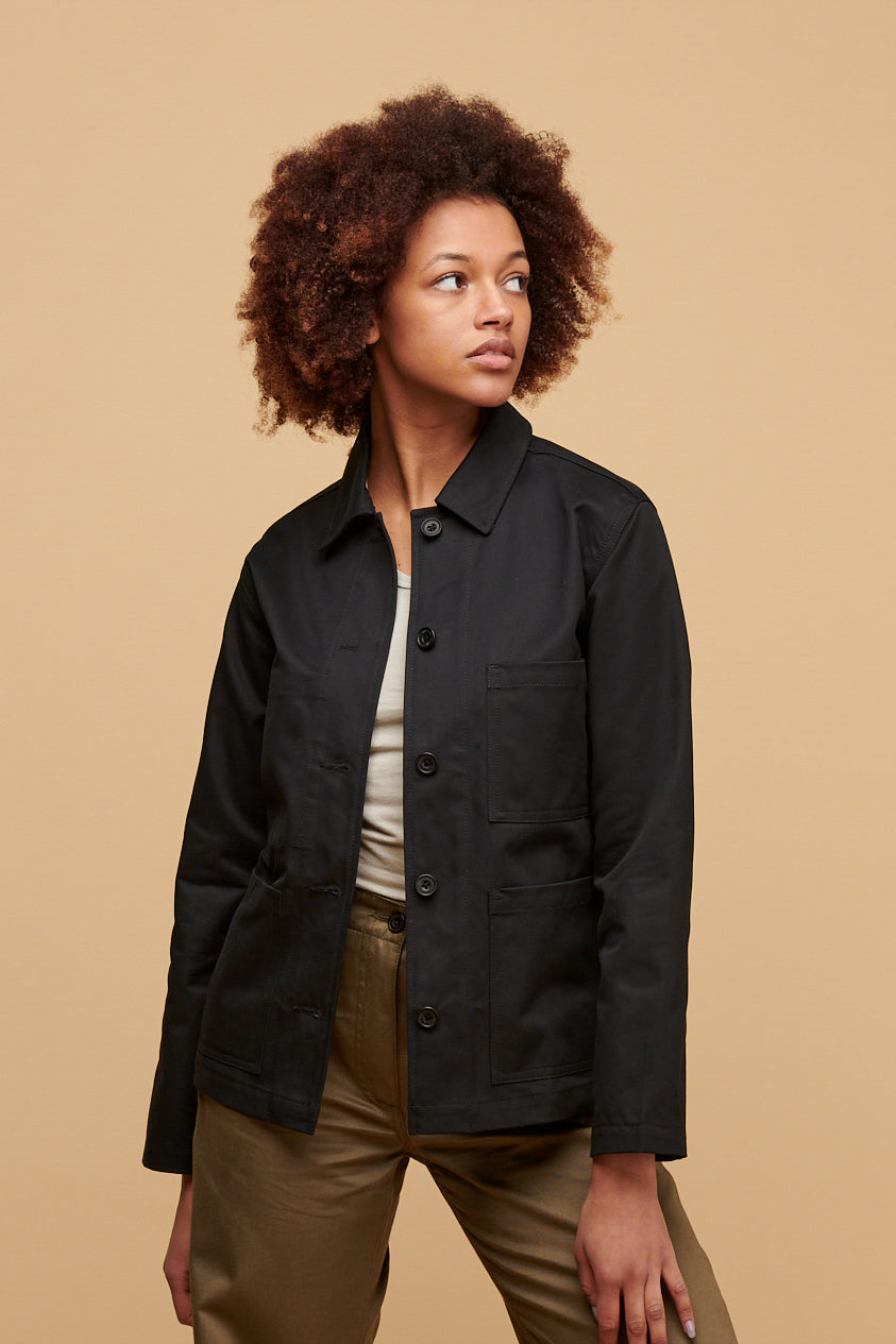 
            Thigh up of female wearing chore jacket in black unbuttoned over racer back vest in stone paired with work trousers in light olive