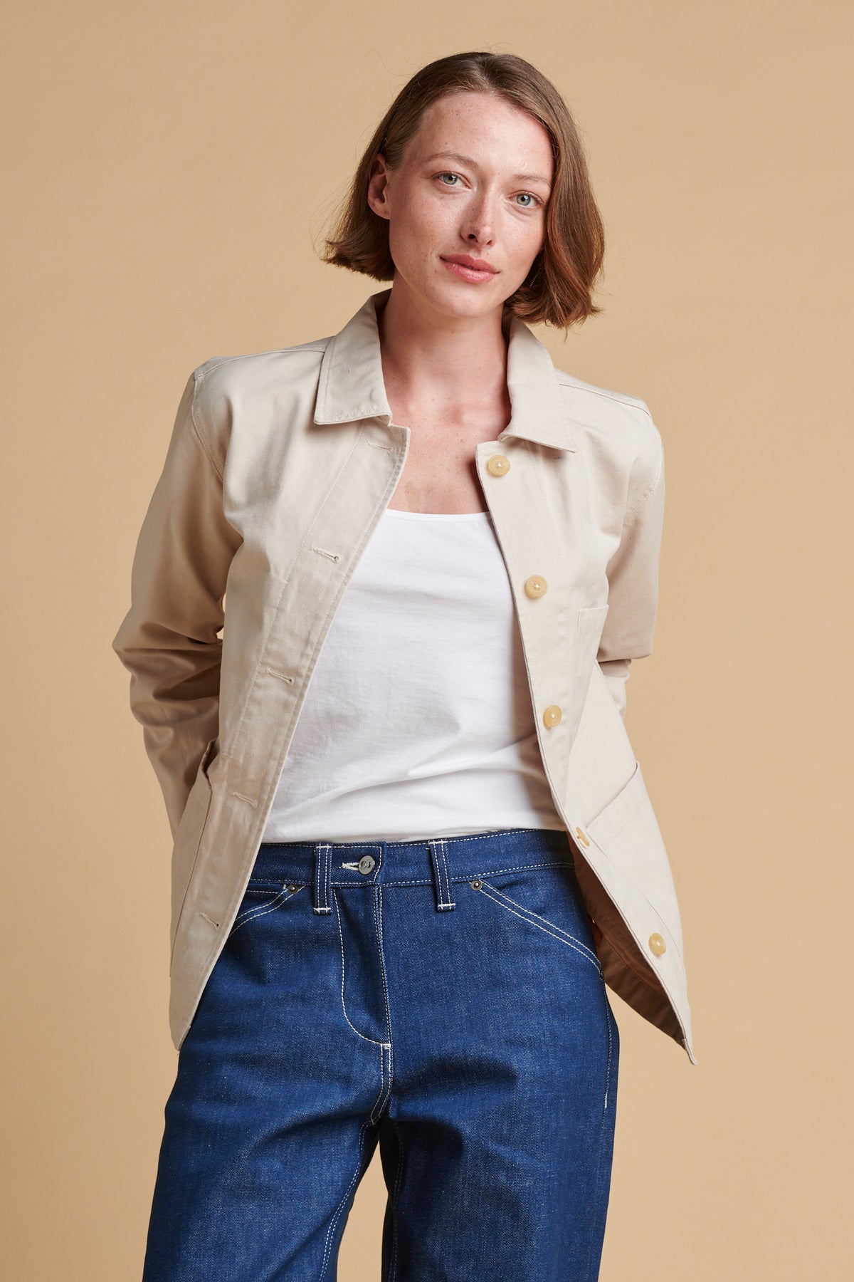 Female wearing chore jacket in cream unbuttoned over white camisole.
