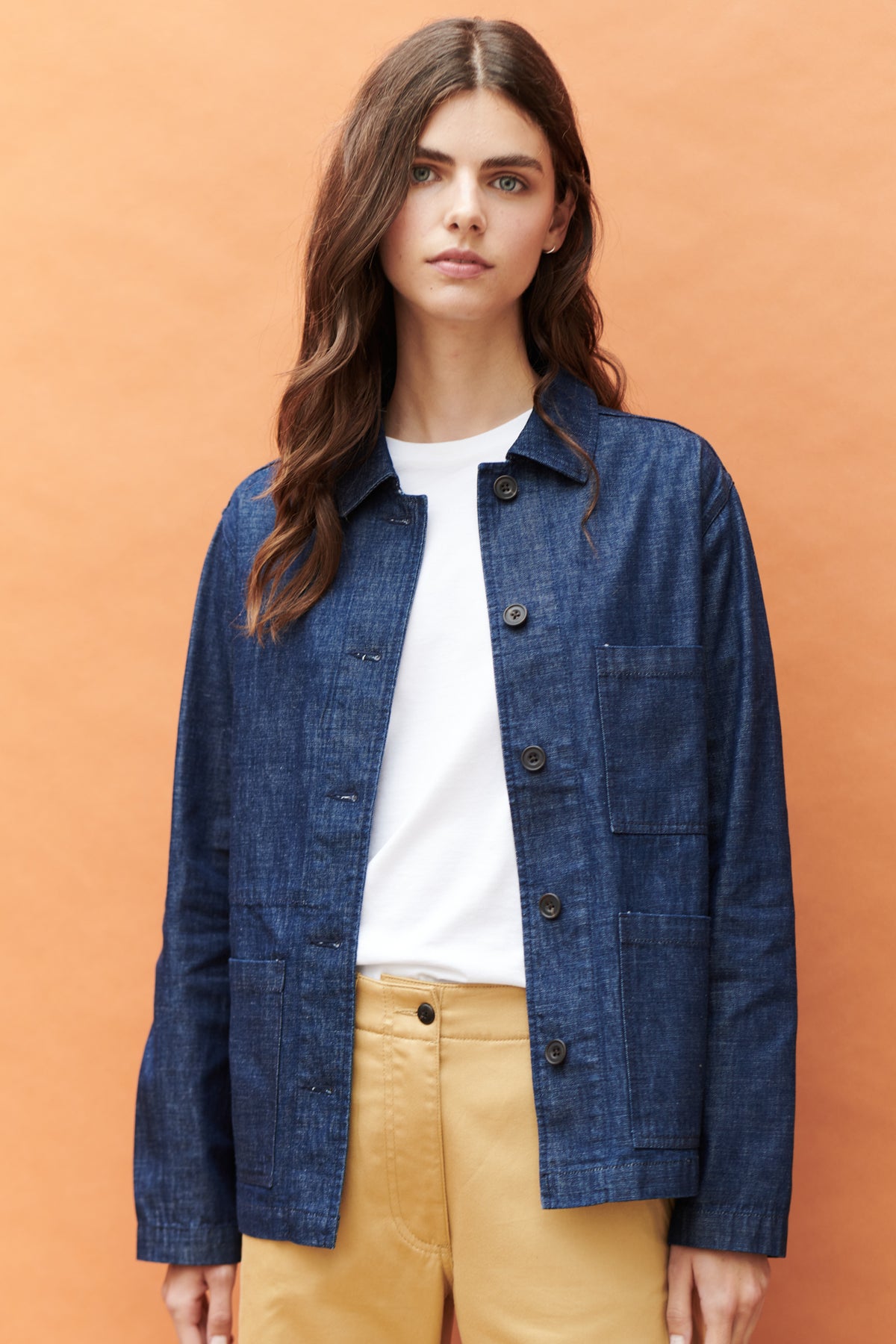 Front, thigh up image of brunette female wearing unbuttoned chore jacket in denim. with three front pockets