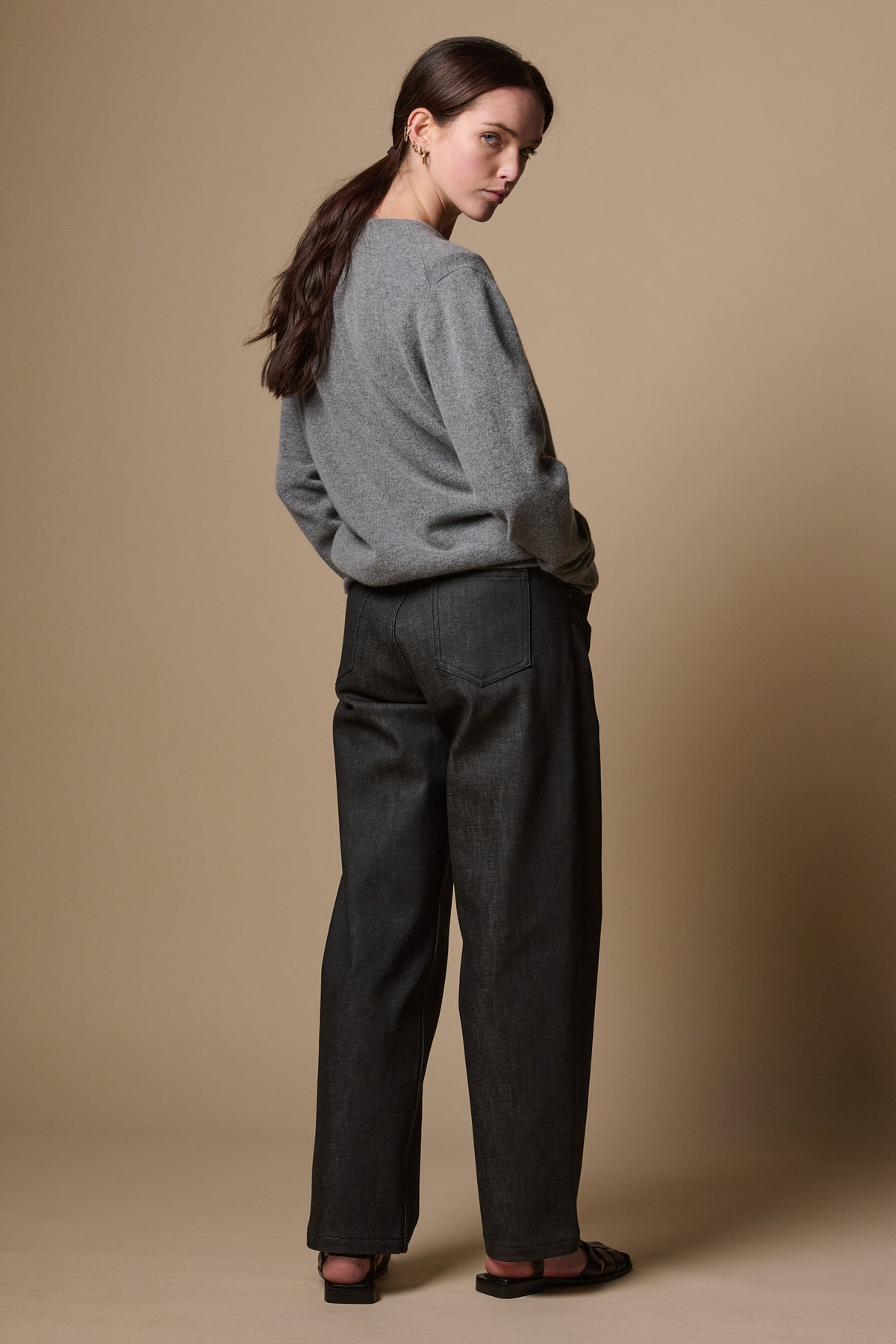 
            The back of white female wearing chore wide tapered leg jeans in black, looking back towards camera with hands in the two front pockets. Worn with lambswool crew neck jumper in grey