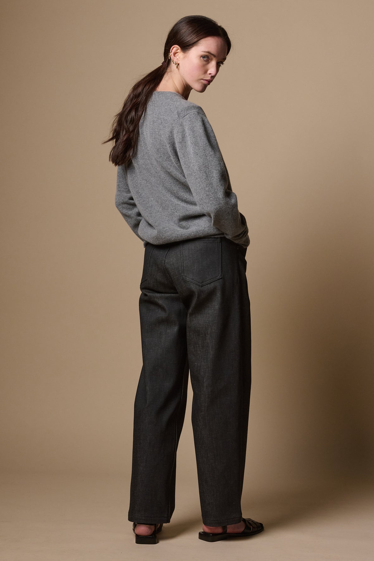 The back of white female wearing chore wide tapered leg jeans in black, looking back towards camera with hands in the two front pockets. Worn with lambswool crew neck jumper in grey