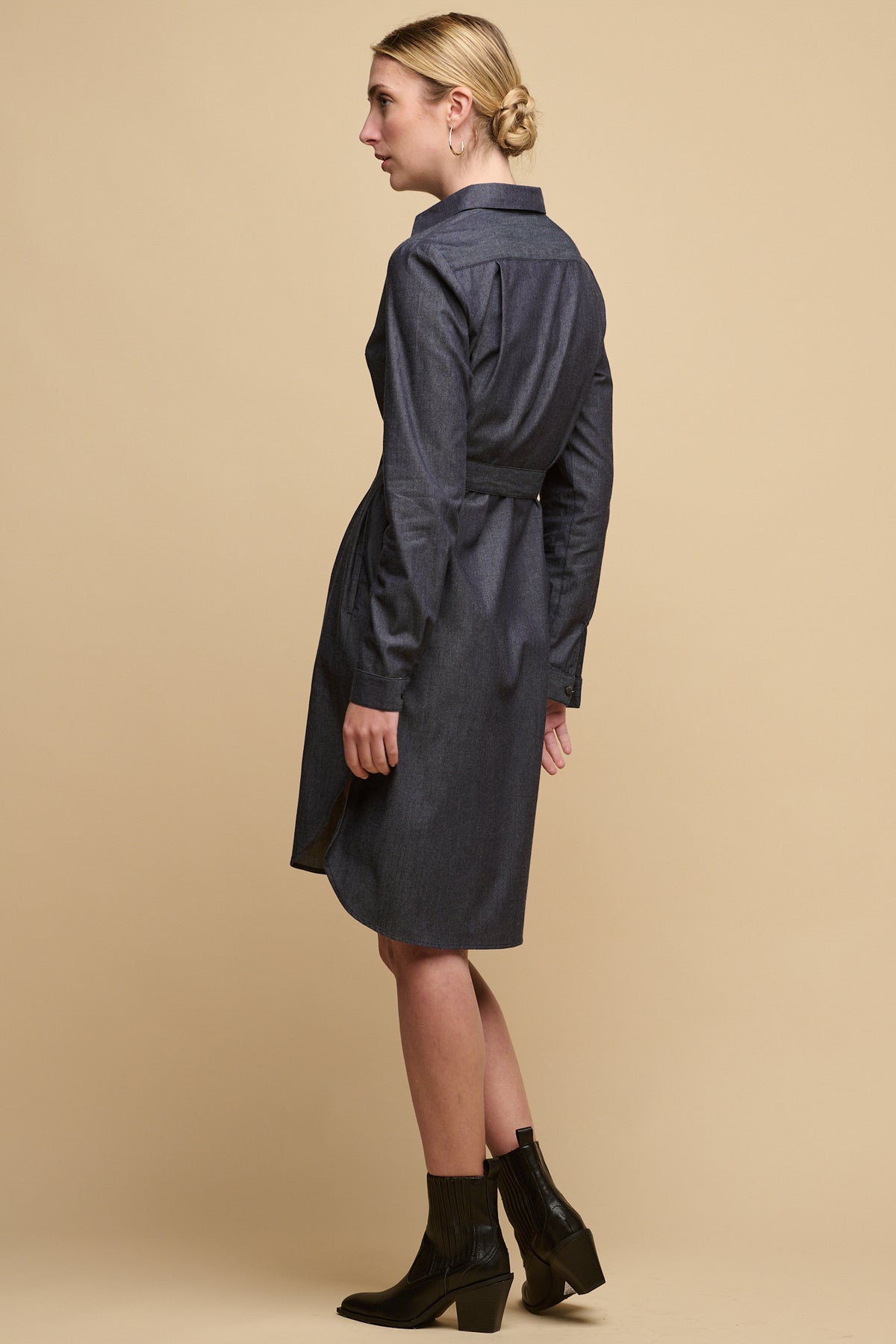 Full body image of the back of female wearing collared shirt dress in indigo paired with heeled boots