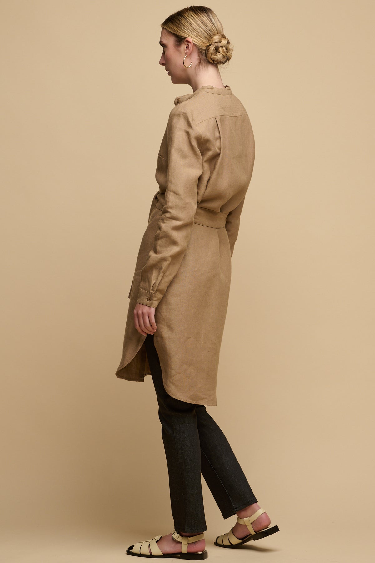 Full body image of the back of female wearing collarless shirt dress in truffle linen with waist belt tie. Dress worn over straight leg jean in black paired with sandals for a day time look.