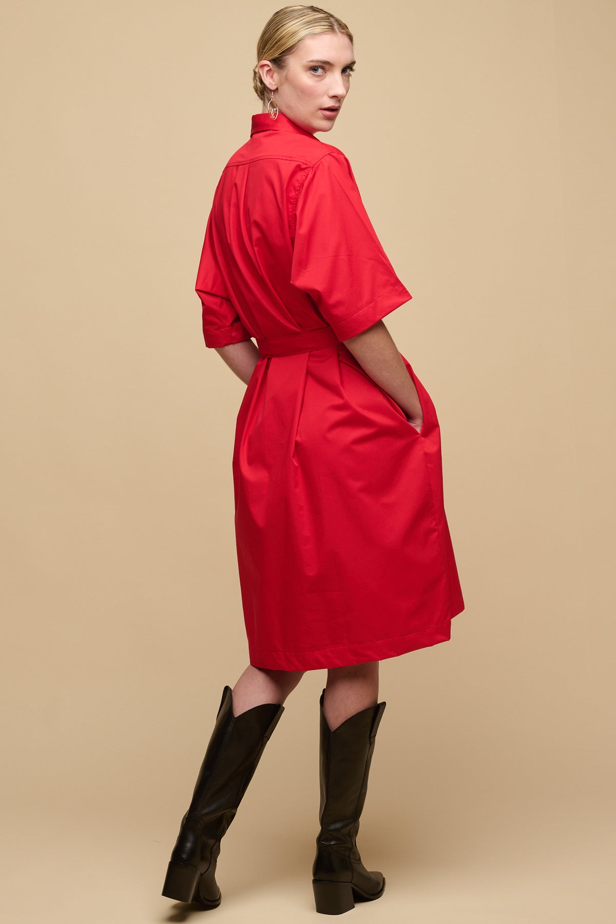 
            Full body back of female wearing cotton dress straight red with waist belt tie and hands in front pockets paired with black boots