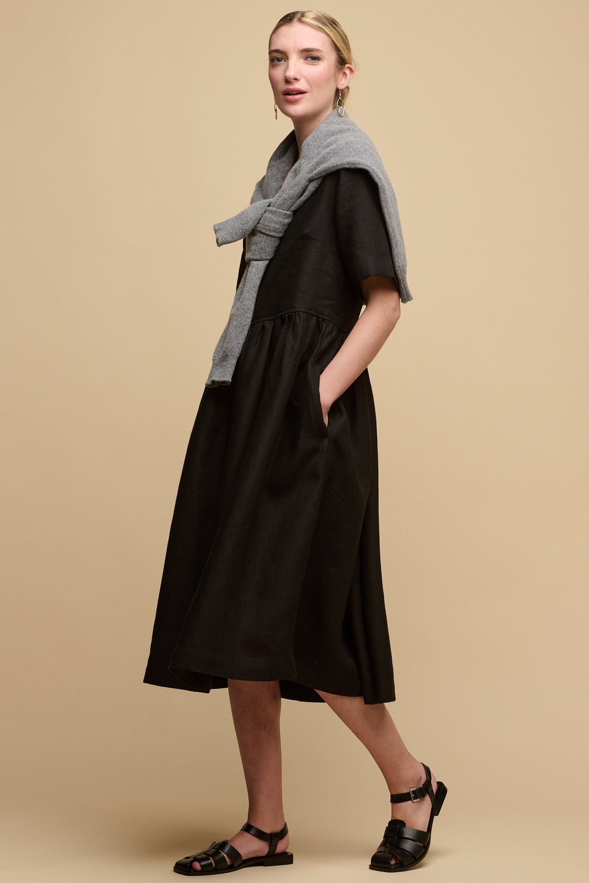 
            Full body image of fair skinned female with hair tied back into a bun wearing crew neck gathered dress in black linen with lambswool jumper draped over the shoulders and paired with sandels.