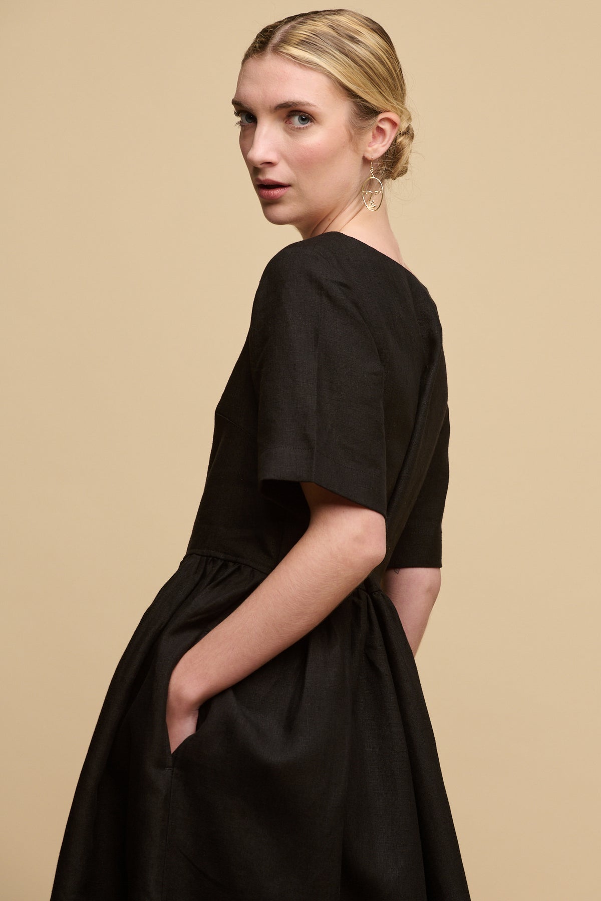 
            Thigh up image of the back of fair skinned female with hair tied back into a bun wearing crew neck gathered dress in black linen with hand in pocket looking back to camera