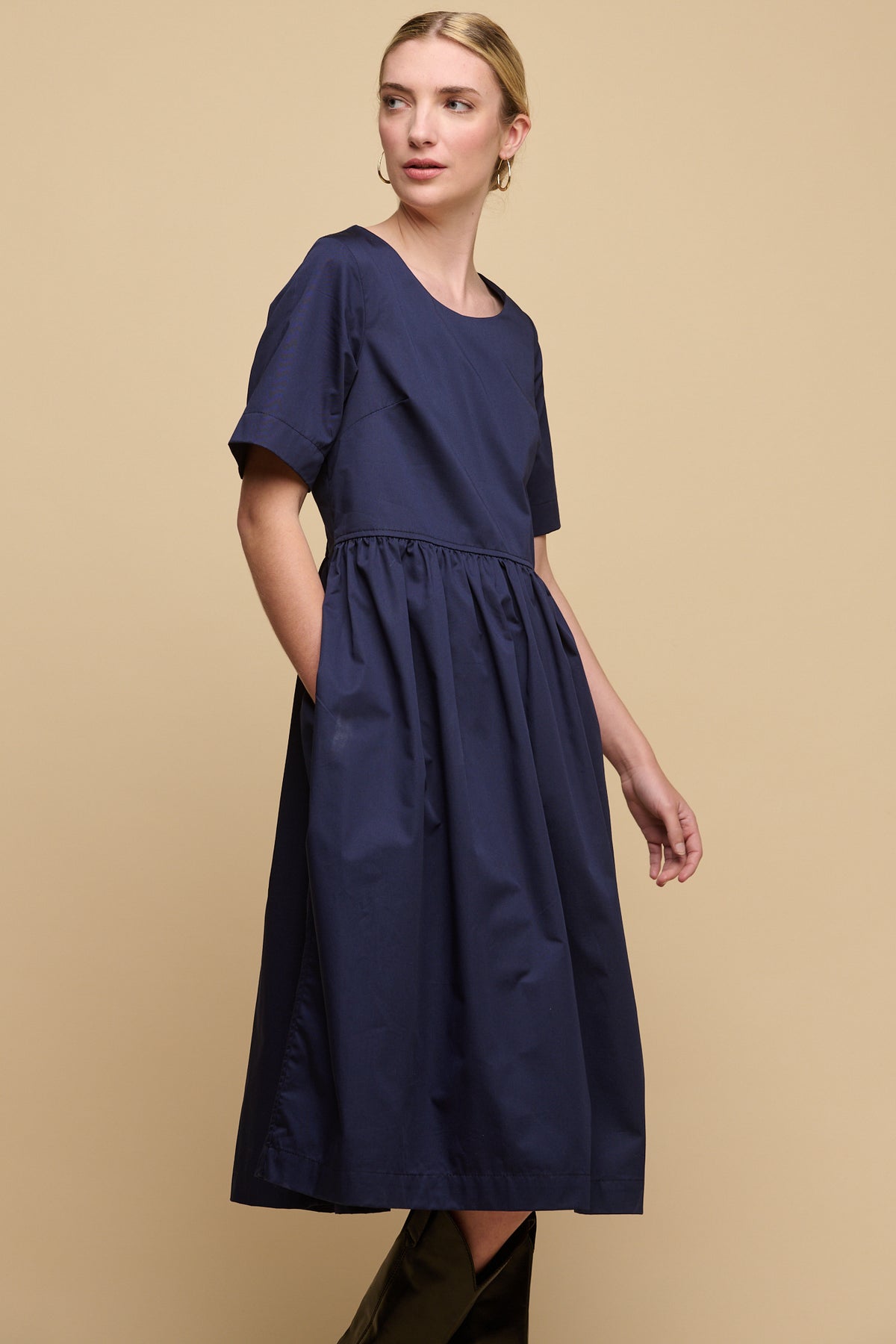 Blonde female with fair skin wearing crew neck gathered dress in navy, with a hand in one of the front pockets