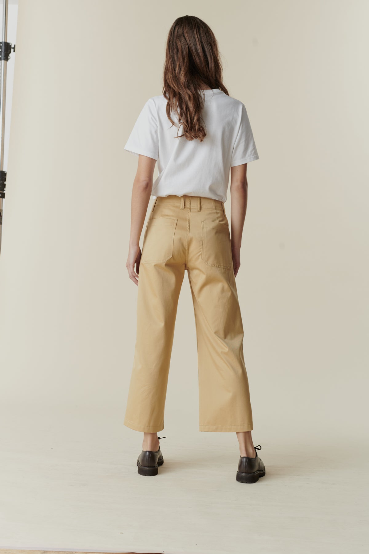 Back, full body image of brunette female wearing cropped work trousers in beige