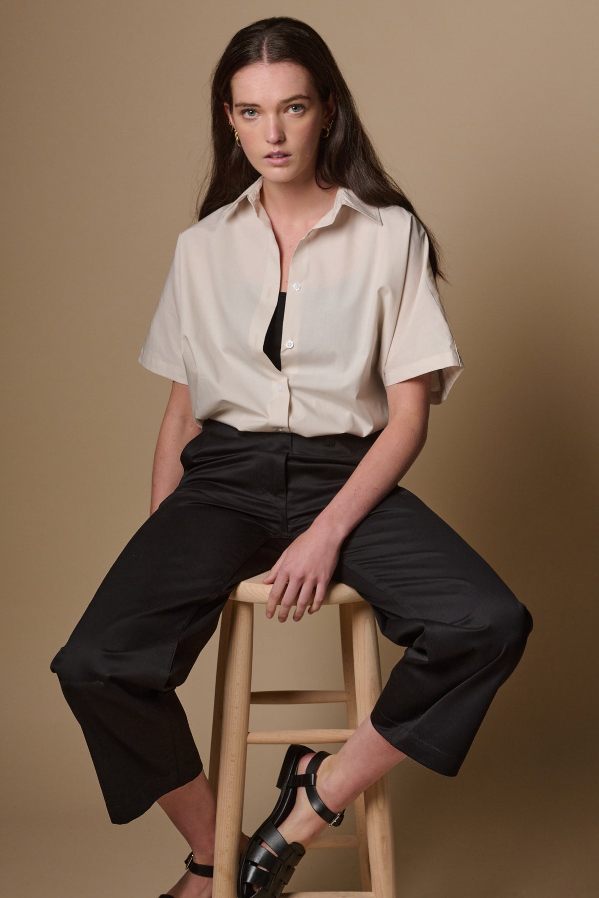 
            Brunette female sat on tall stool wearing cropped work trousers in black paired with ava short sleeve shirt in stone