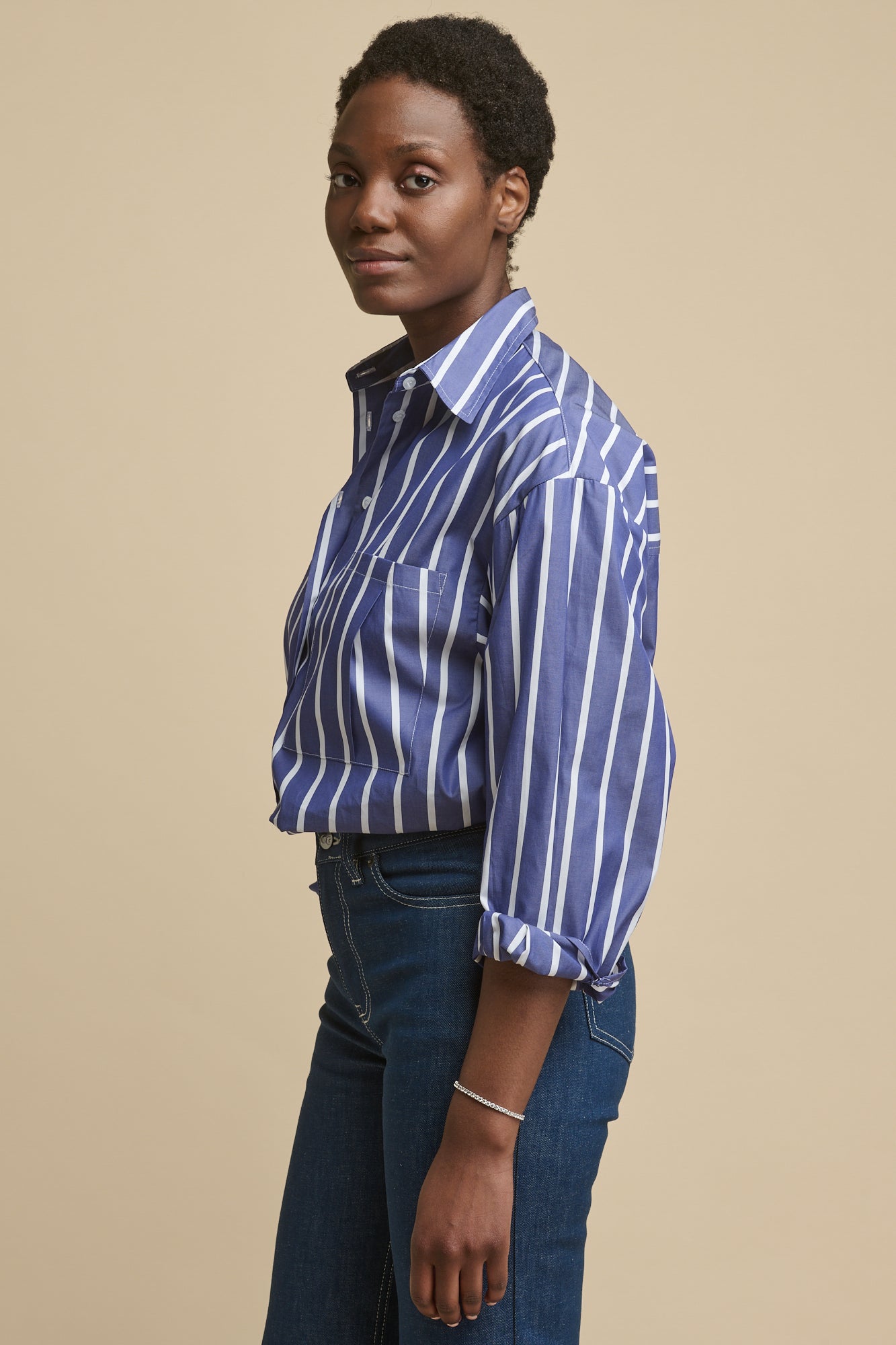 Thigh up image of the side of female wearing long sleeve Esme shirt in blue and white stripe tucked into blue jeans. Pleat detail patch pocket on chest and sleeves rolled up
