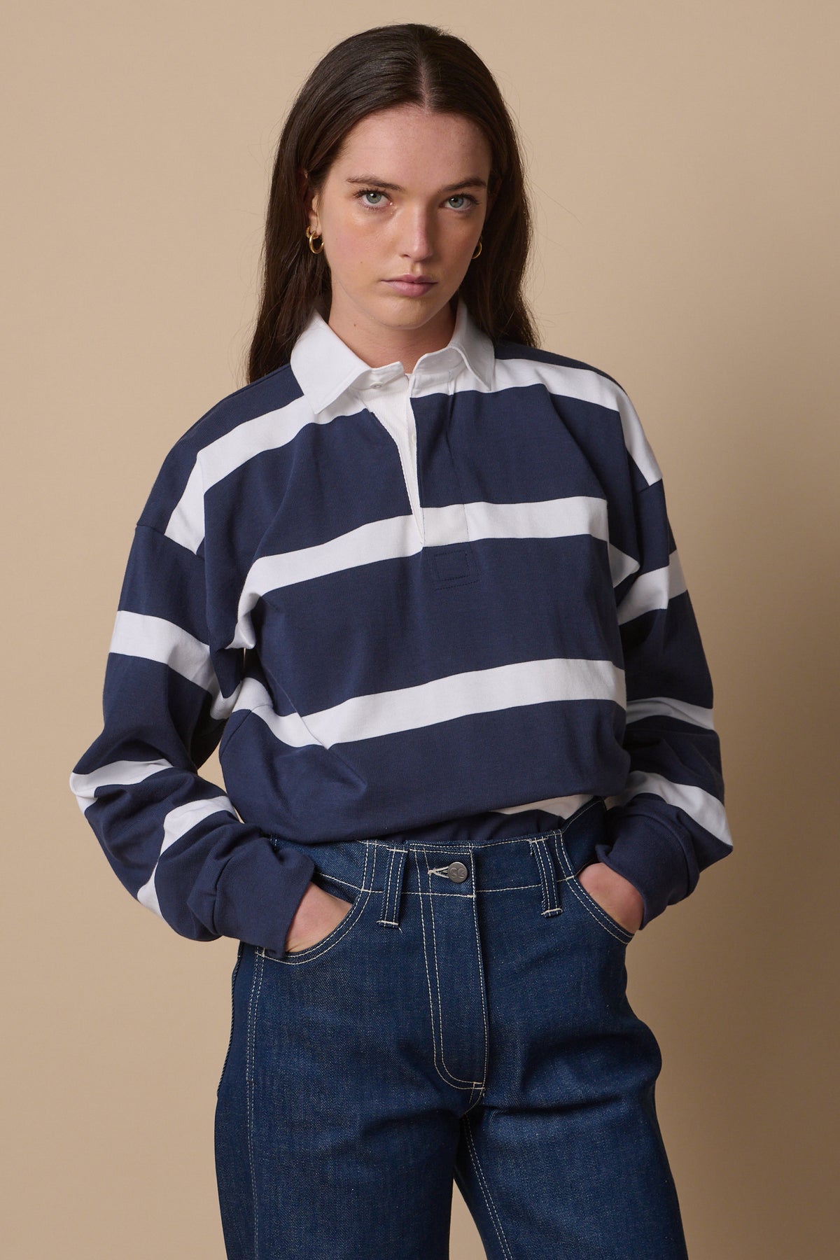 Thigh up image of brunette female wearing unisex fine stripe rugby shirt tucked into work jeans in blue