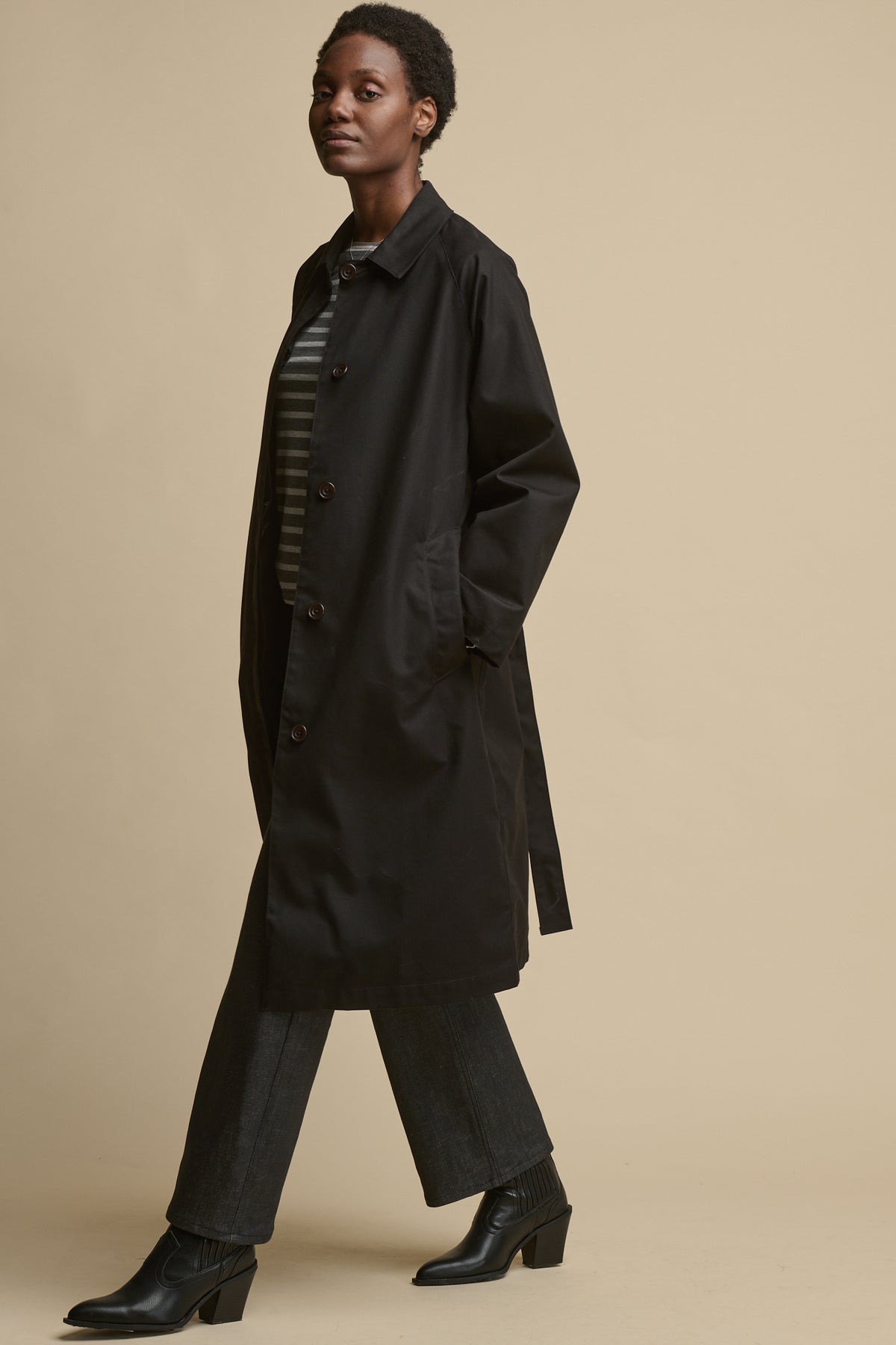 Full body image of female wearing unfastened women's Frances Raglan Belted Raincoat in black with her hands in the front two pockets
