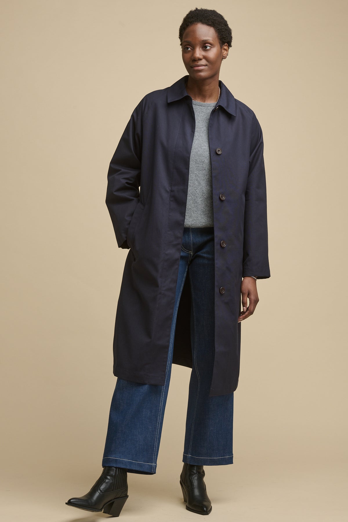 
            Full body image of female wearing women&#39;s Frances Raglan Belted Raincoat in navy unfastened over grey lamsbwool crew neck jumper and blue jeans