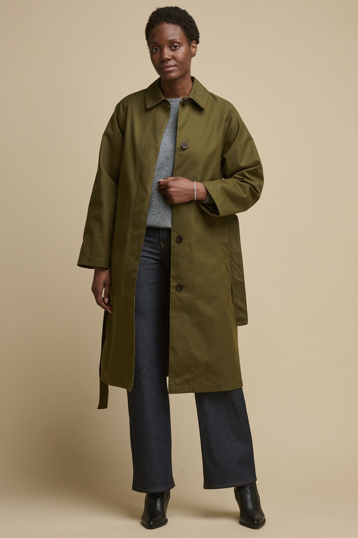 Full body image of female wearing Frances Raglan Belted raincoat in olive unfastened over lambswool crew neck jumper with indigo jeans