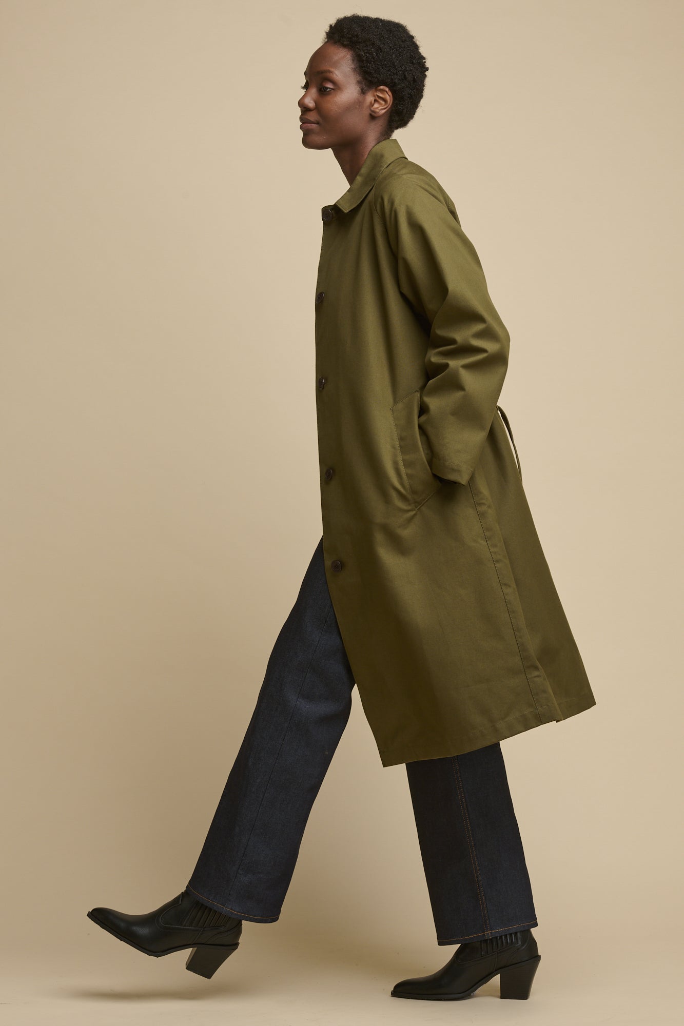 Full body image of female from the side, mid walk wearing Frances Raglan Belted Raincoat in olive with indigo jeans and black boots