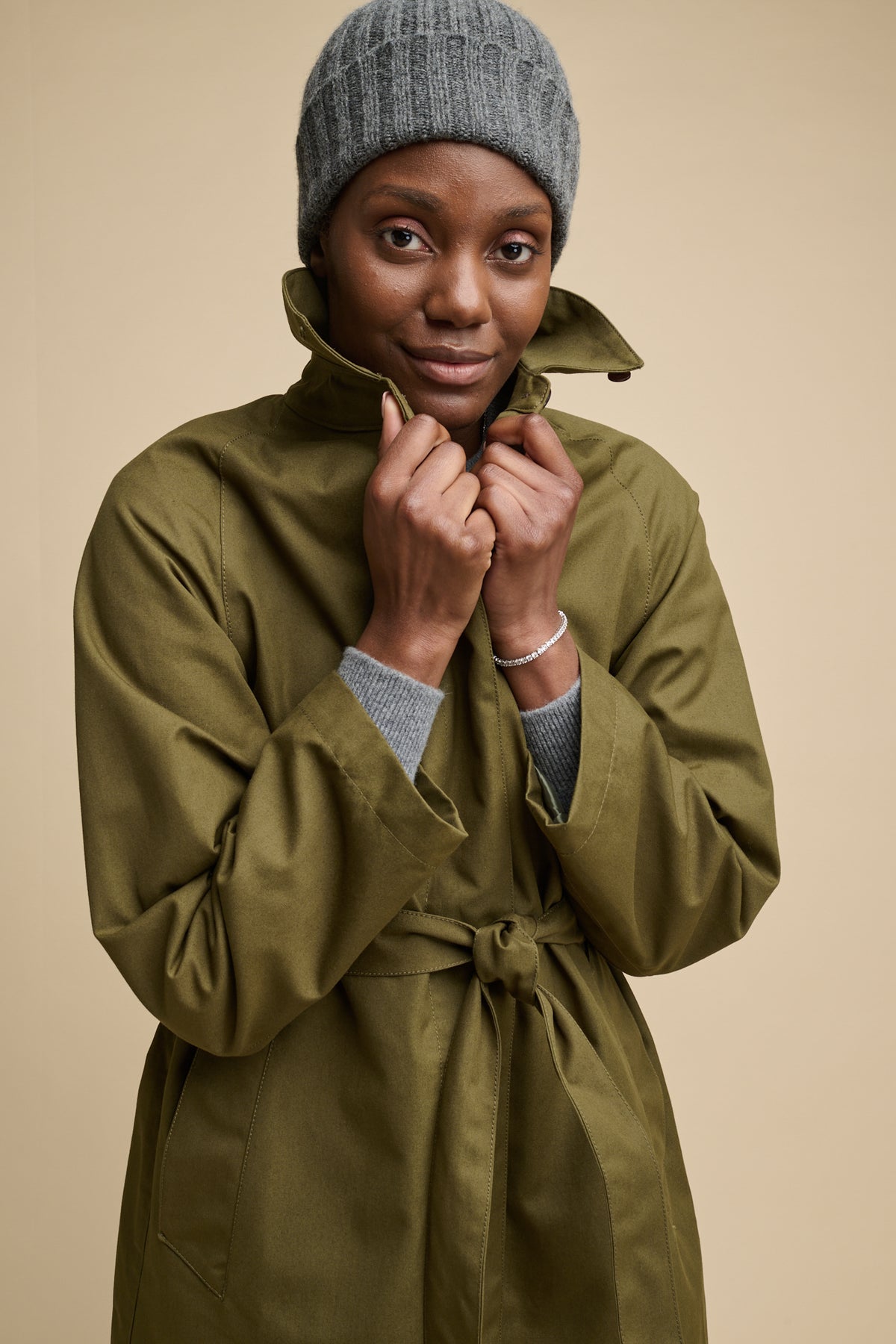 
            Thigh up image of the front of female wearing Frances Raglan raincoat in olive belted and fastened with the collar up, worn with beanie hat in grey