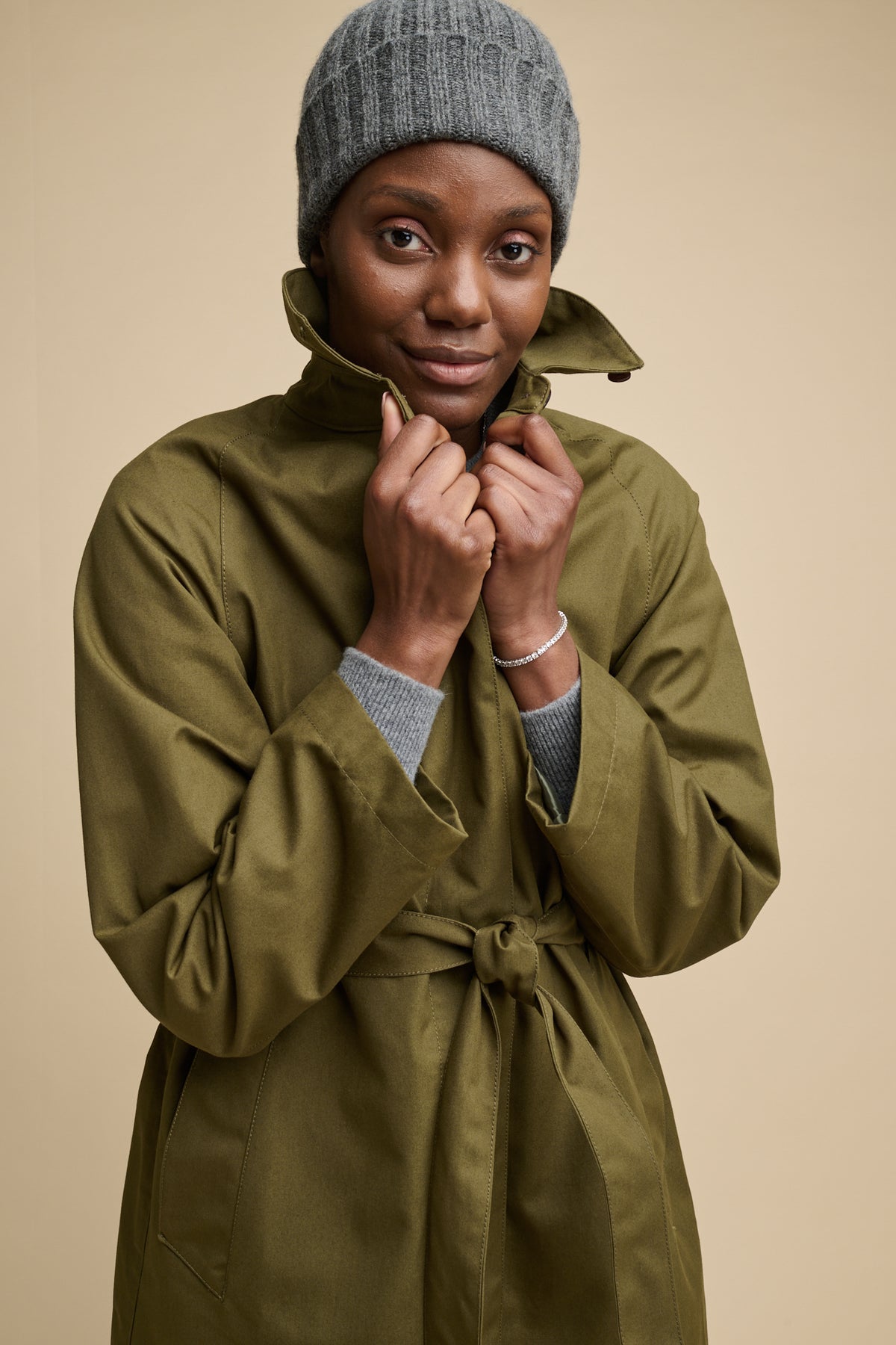 Thigh up image of the front of female wearing Frances Raglan raincoat in olive belted and fastened with the collar up, worn with beanie hat in grey
