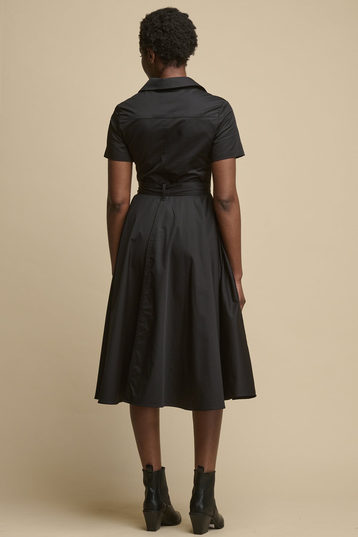 
            Full body image of the back of black female wearing full skirted phoebe trench dress in black, paired with black boots