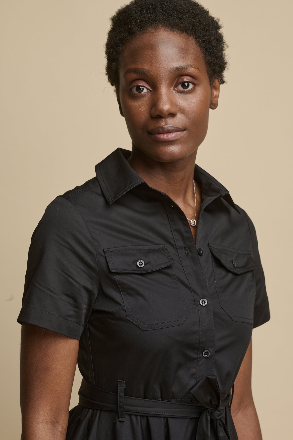 
            Close up portrait of female wearing full skirted phoebe trench dress in black, with short sleeves and two front chest pockets