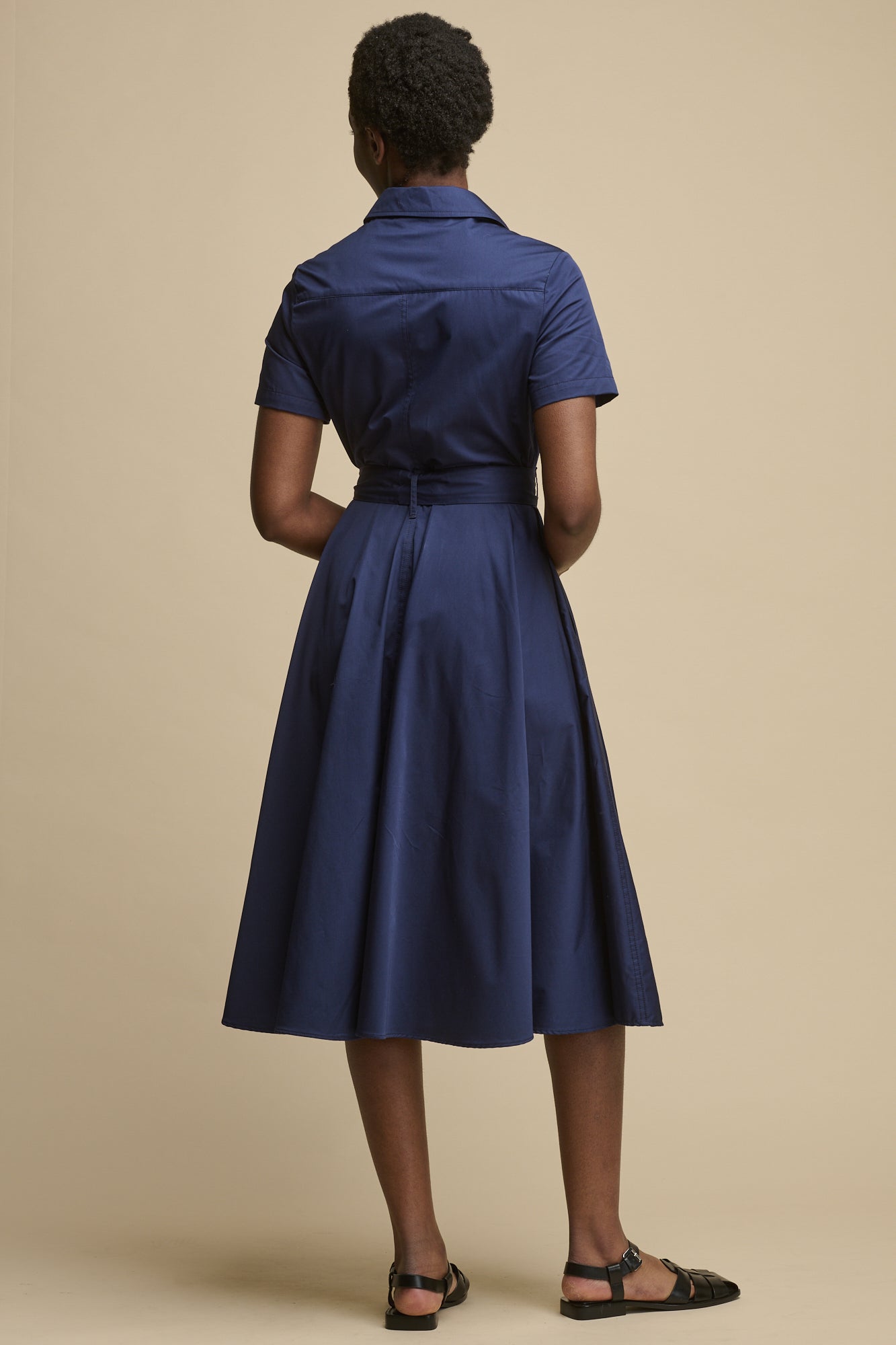 Full body image of female from the back wearing full skirted phoebe trench dress in navy with short sleeves and belt tie, midi length dress paired with black sandals
