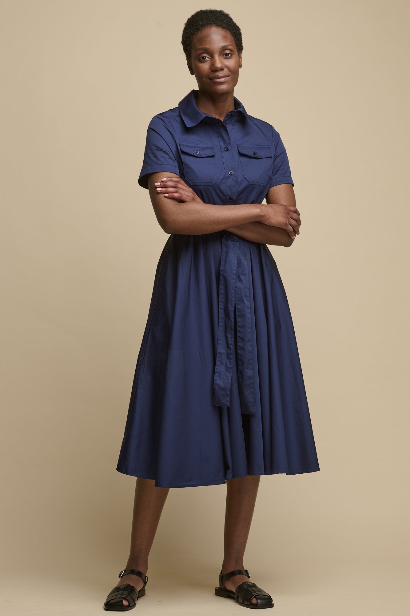 Full body image of the front of female wearing short sleeve shirt dress, with her arms folded over the waist belt tie, midi length dress