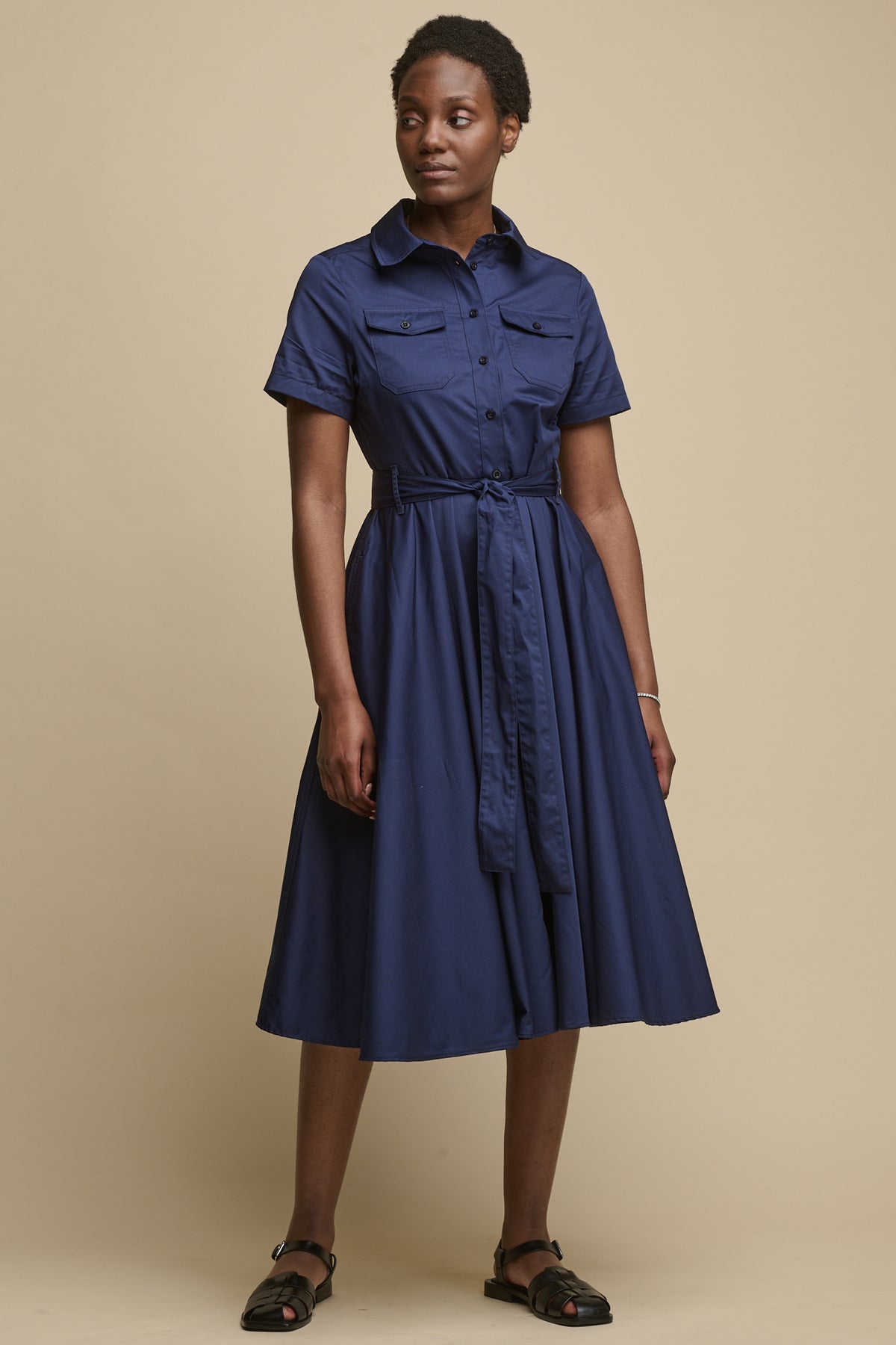 Full body image of female wearing full skirted phoebe trench dress in navy with belt tied around waist, two front chest pockets.