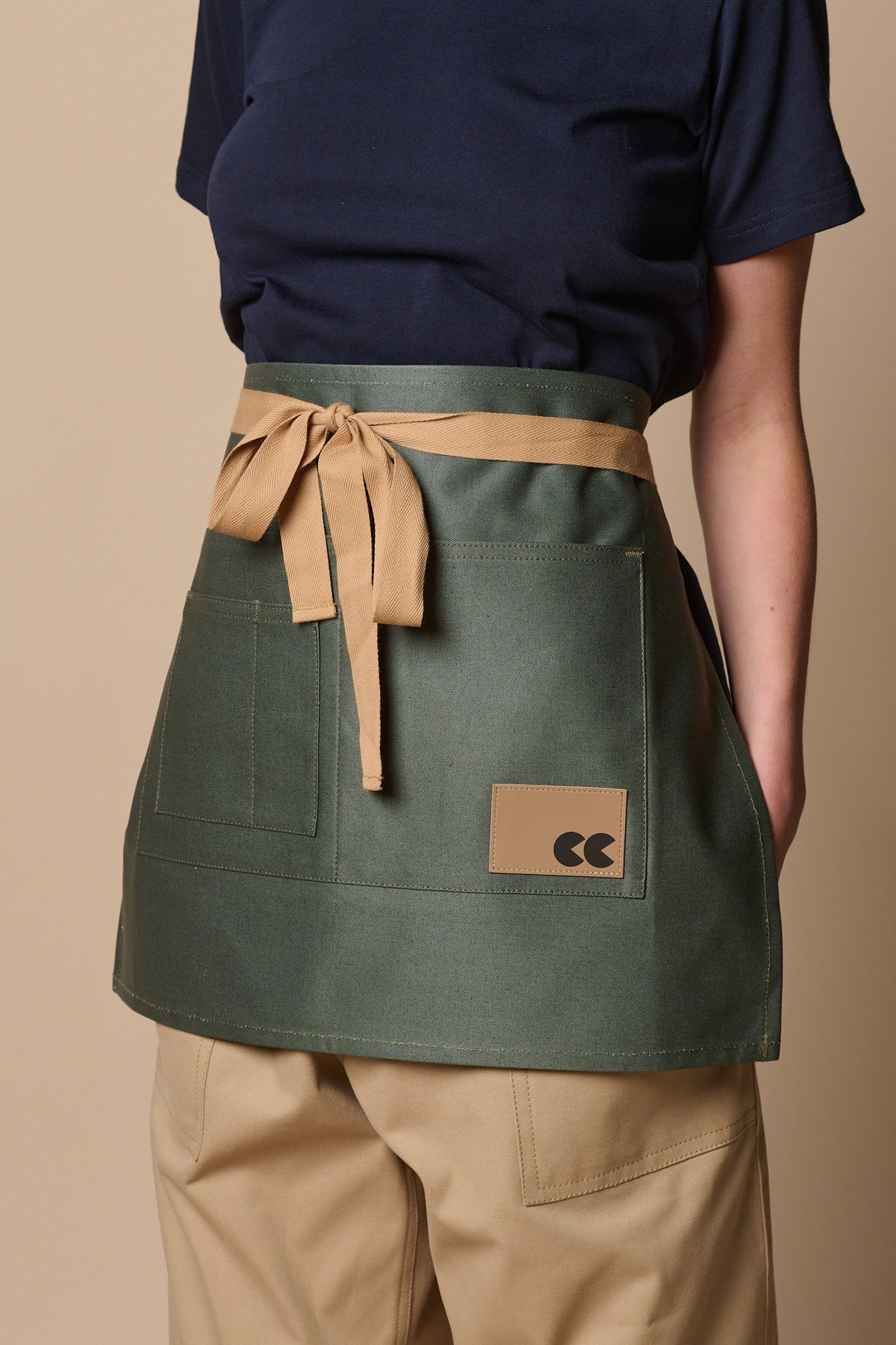 
            Female wearing garden apron in forest, with beige ties tied in a bow at the front. Two large and two smaller pockets on the front of apron. CC logo patch