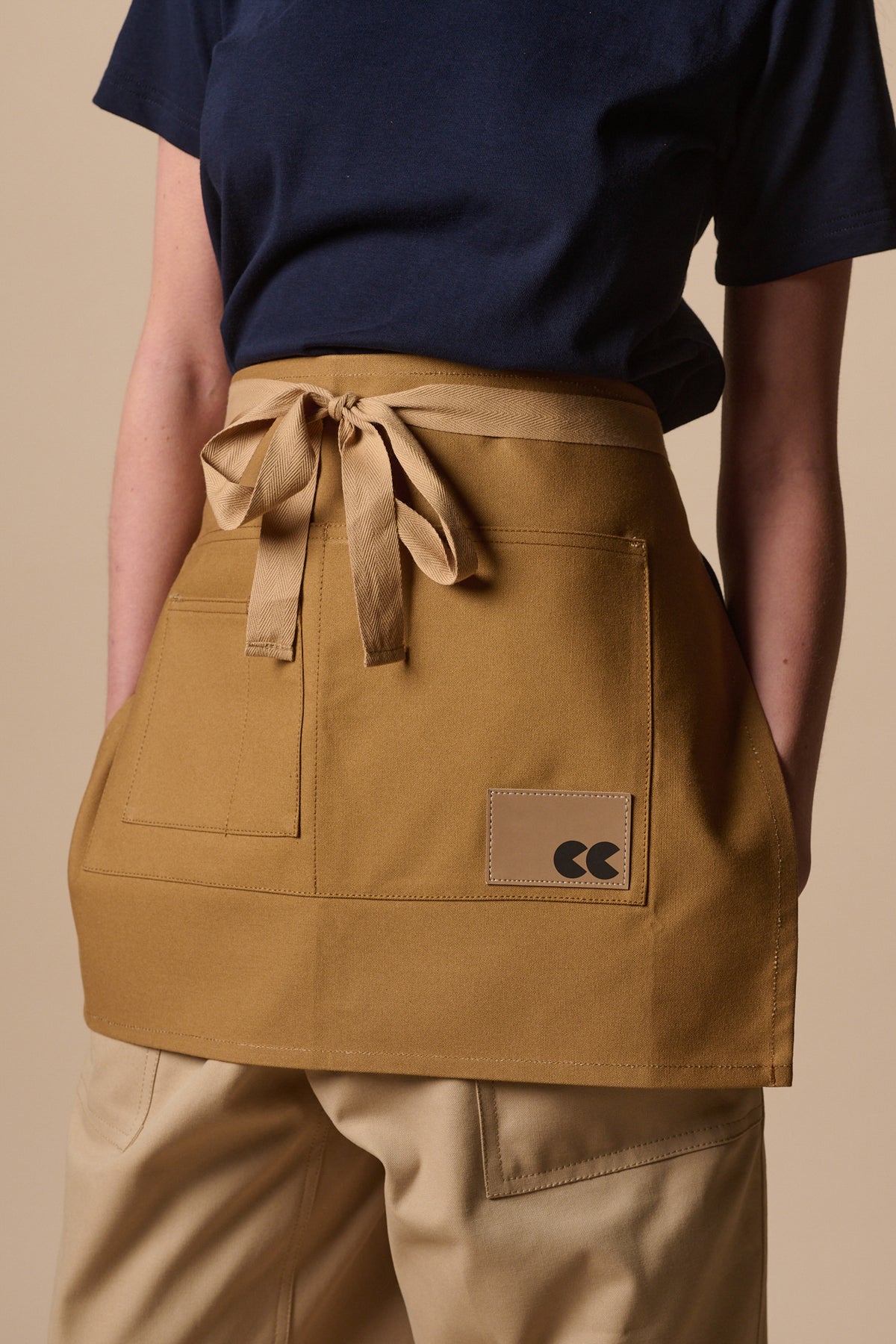 
            Female wearing unisex garden apron in khaki with front pockets and CC logo badge.