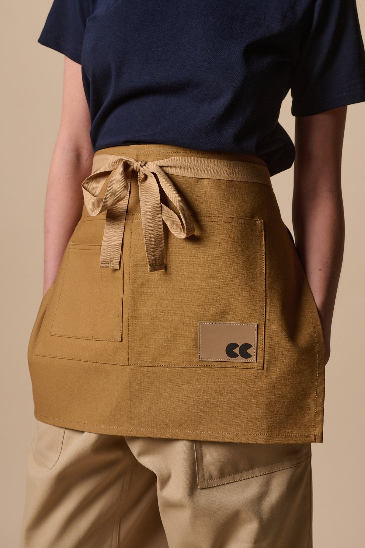 Female wearing unisex garden apron in khaki with front pockets and CC logo badge.