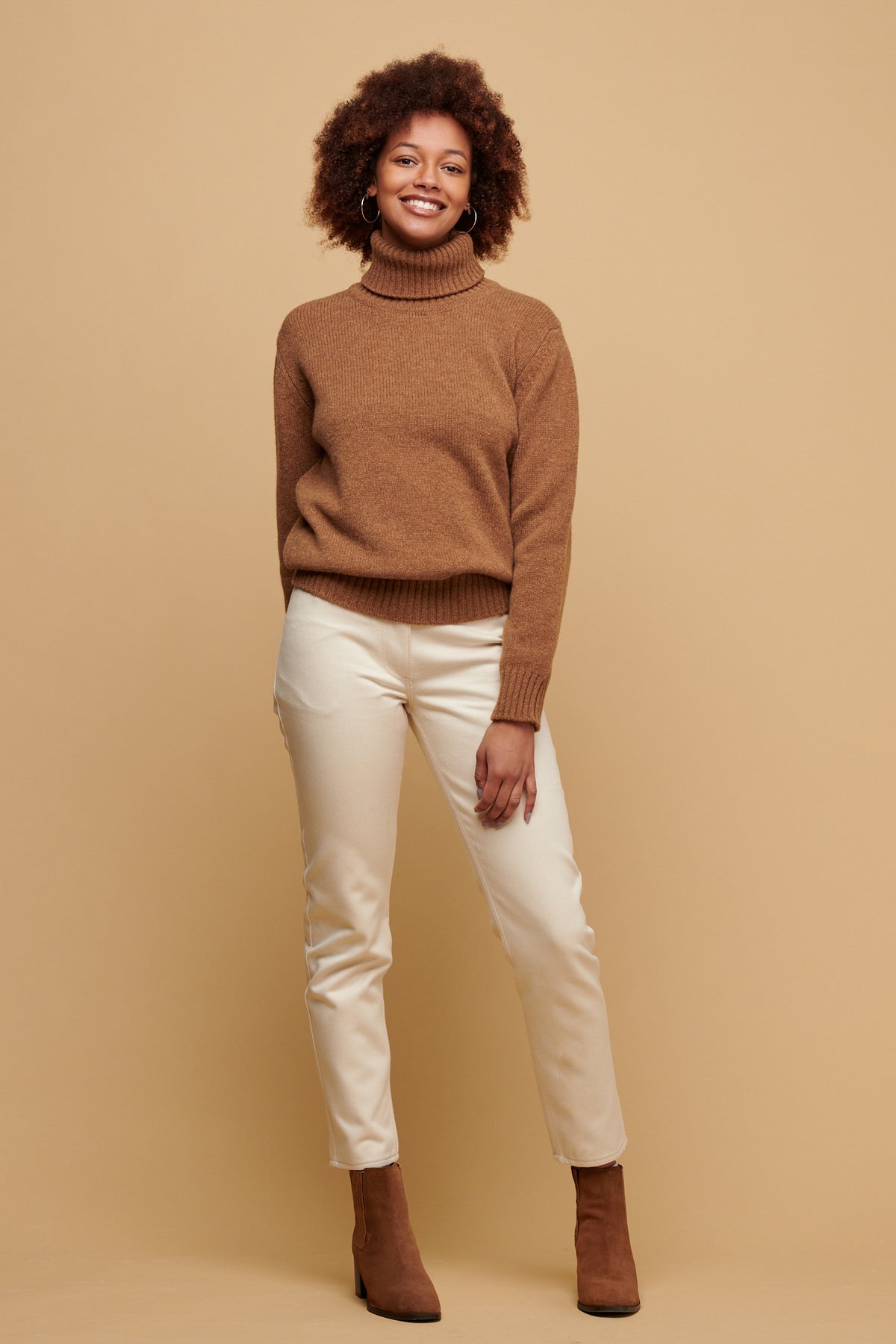 The front of female of colour with an afro wearing lambswool roll neck jumper in dark camel paired with Gloria jeans in ecru and a dark camel coloured suede boot