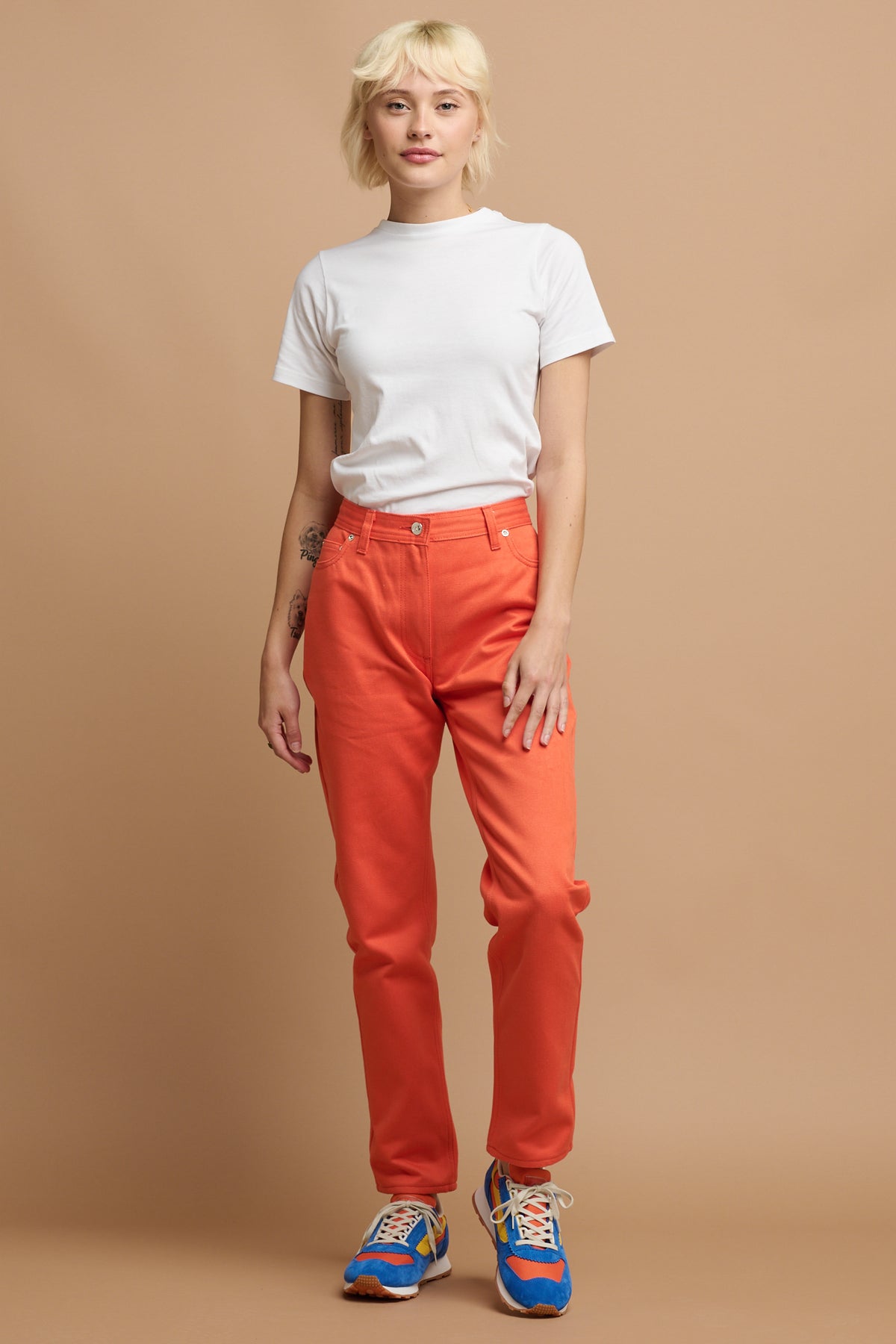 Female with short blonde hair wearing Community Clothing Gloria Tapered Jean in flame paired with white t shirt tucked in and Parlick trainers