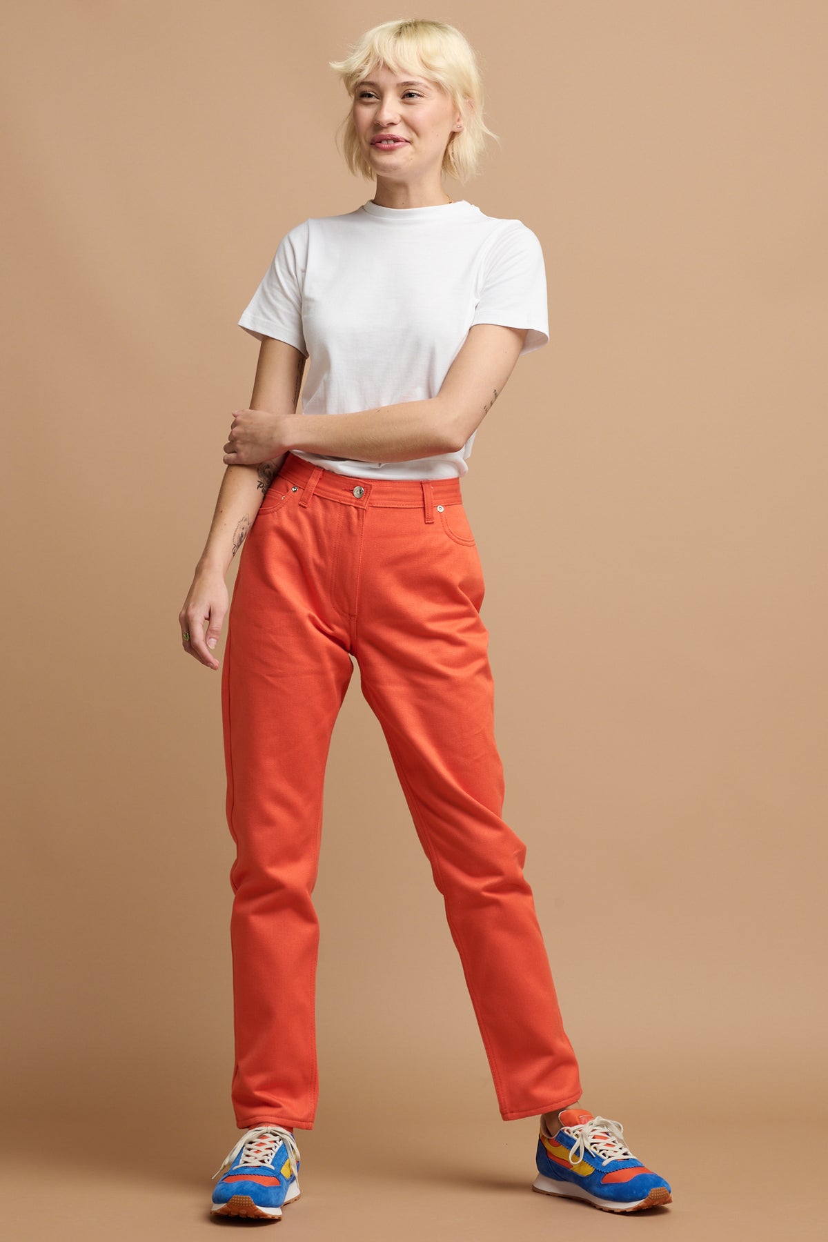 Smiley female with short blonde hair wearing Community Clothing Gloria Tapered Jean in flame paired with white t shirt tucked in and Parlick trainers
