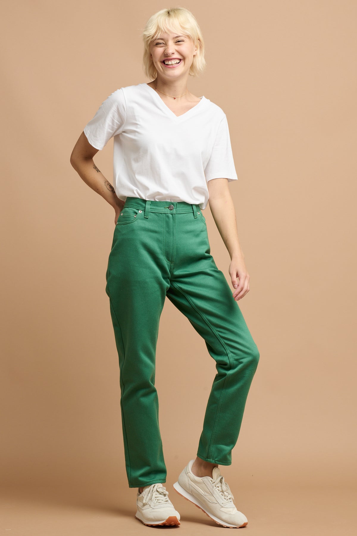 
            Smiley blonde female wearing Community Clothing Gloria Tapered Jean in green with v neck white t shirt and ivory Parlick trainers