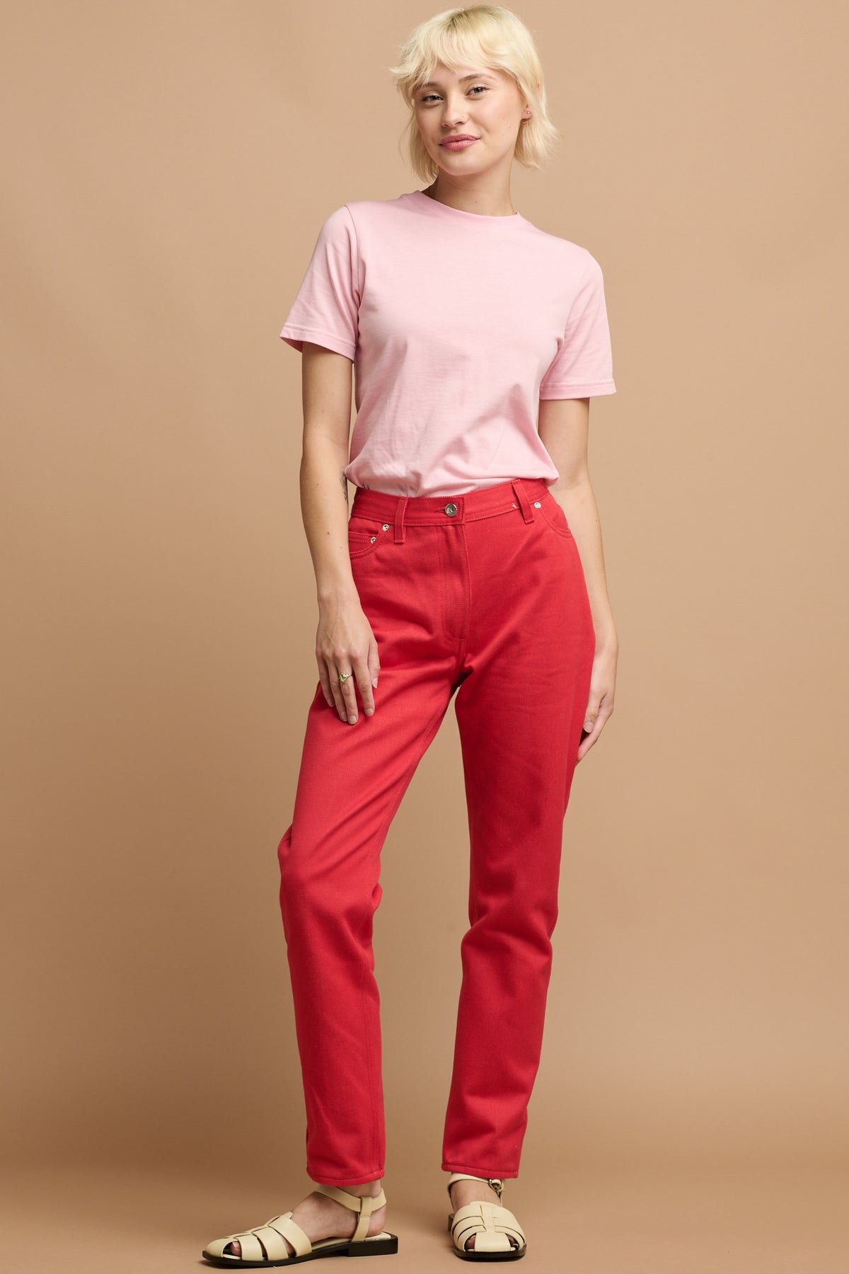 
            White female with short blonde hair wearing Community Clothing Gloria tapered jeans in poppy red paired with short sleeve t shirt in pink tucked in and cream sandals