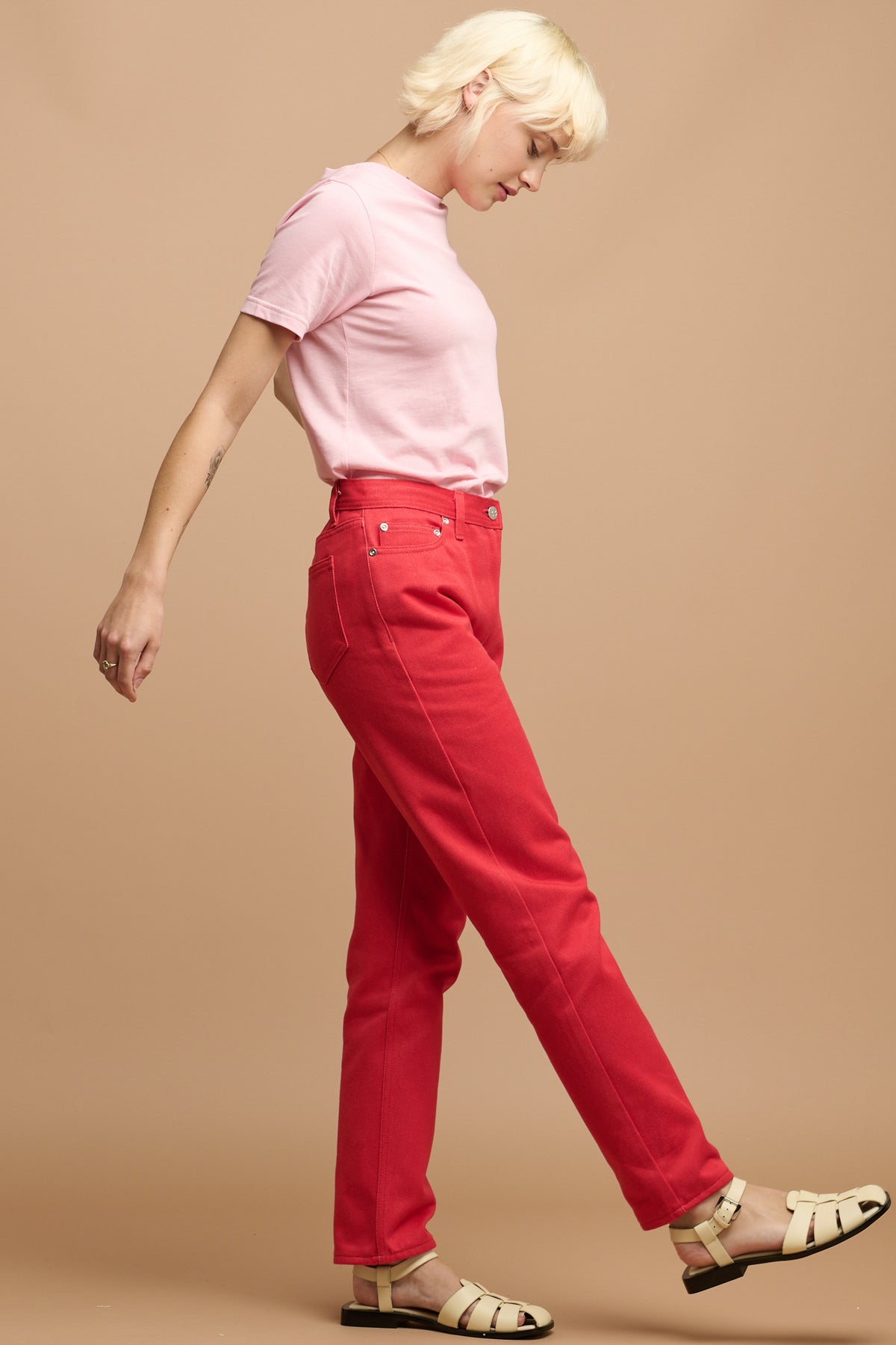 
            The side of white female with short blonde hair wearing Community Clothing Gloria tapered jeans in poppy red paired with short sleeve t shirt in pink tucked in and cream sandals