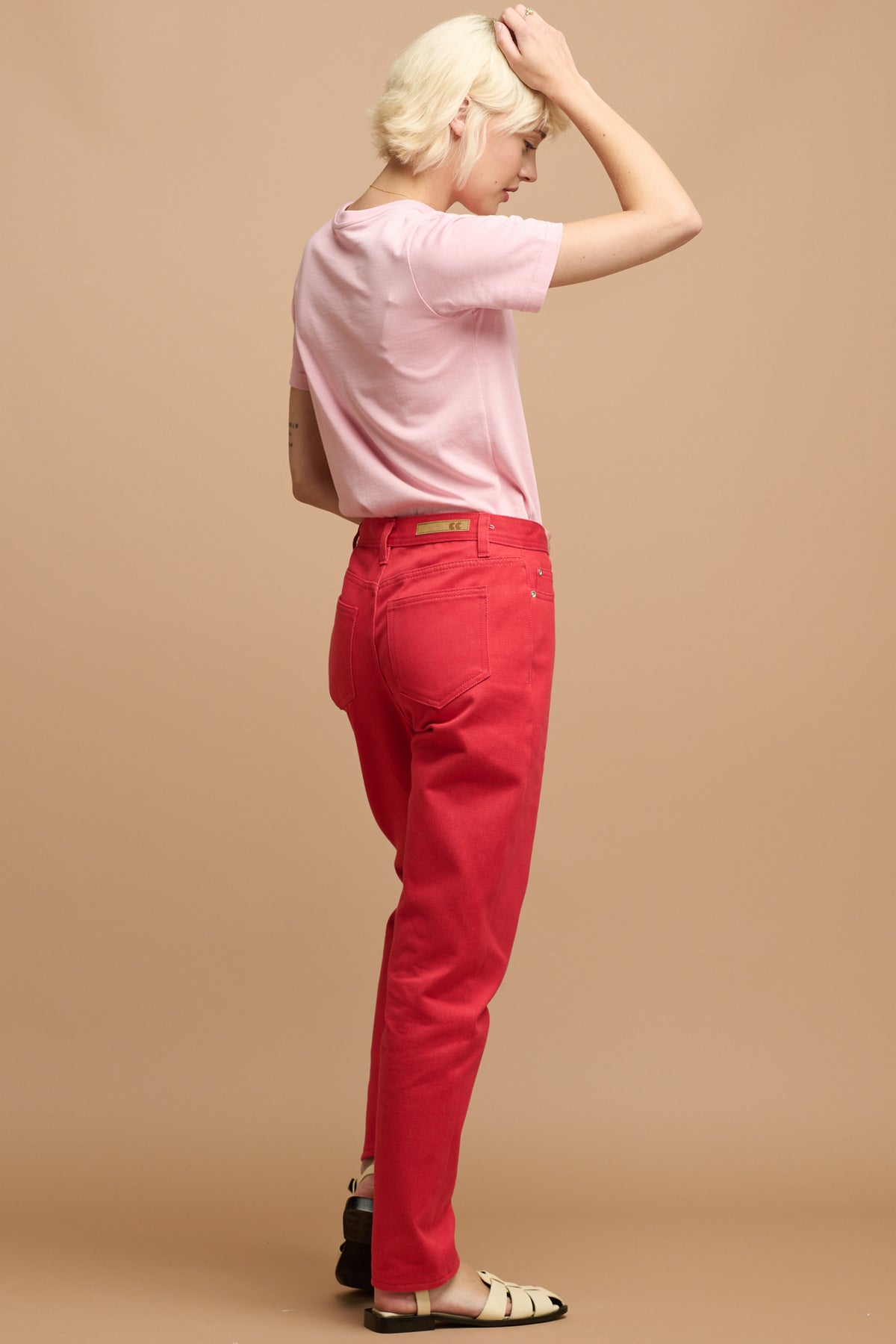 
            the back of white female with short blonde hair wearing Community Clothing Gloria tapered jeans in poppy red paired with short sleeve t shirt in pink tucked in and cream sandals