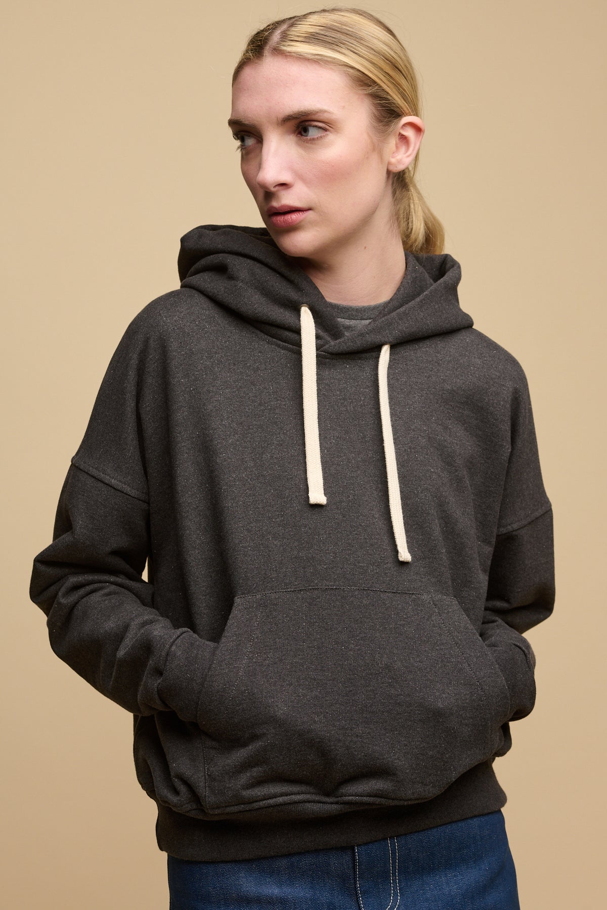 Blonde female wearing heritage hooded sweatshirt in charcoal with hands in front pocket worn over crew neck t shirt in charcoal. Ecru drawstring through the hood