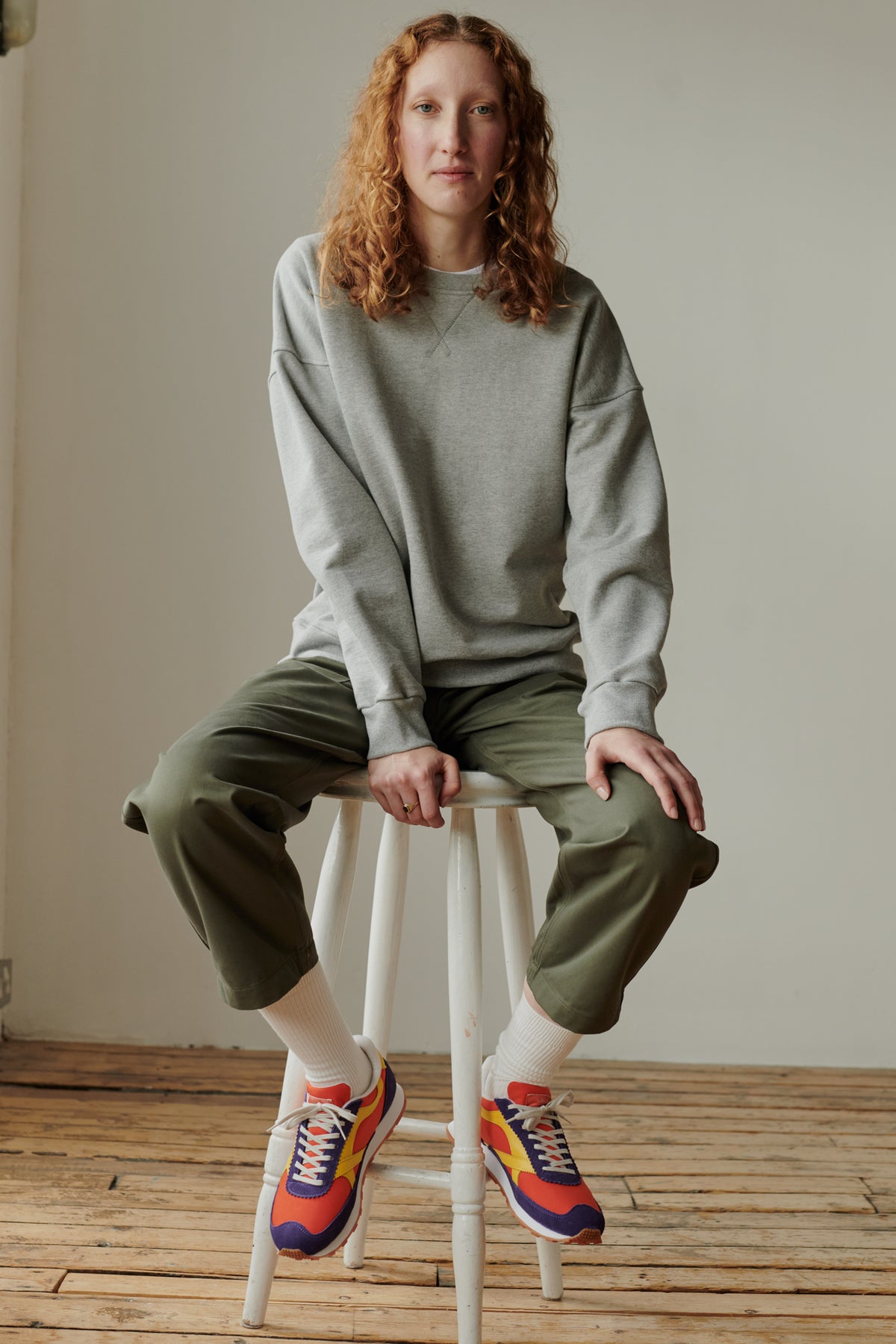 
            Curly haired ginger female wearing women&#39;s heritage sweatshirt in grey face on sat on tall stool 