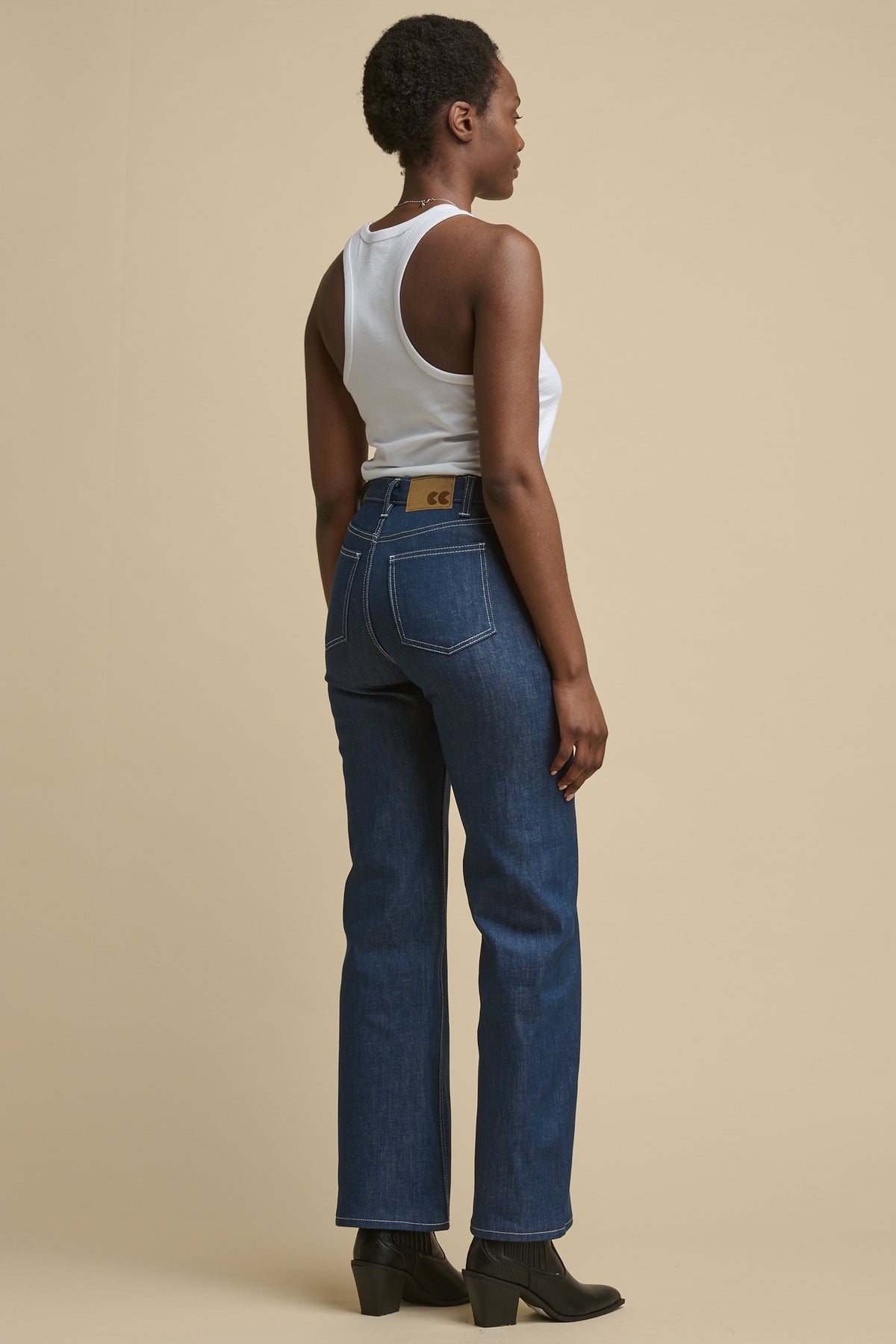 
            full body image of the back of a black woman wearing community clothing high rise wide leg jeans in blue with CC logo jeans patch on the back of waist band, with white contrast stitching. Worn with white vest and black boots