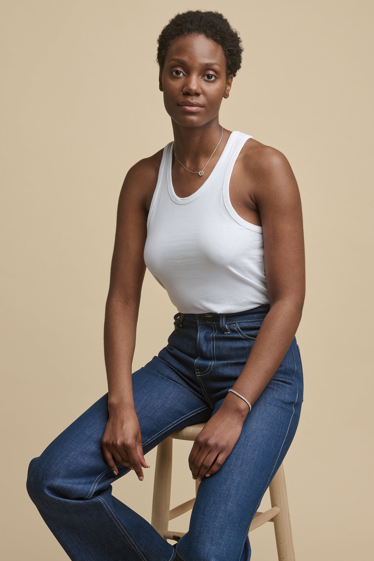 
            Thigh up image of female sat on wooden stool wearing high rise wide leg jeans in blue with white contrast  paired with white racer back vest