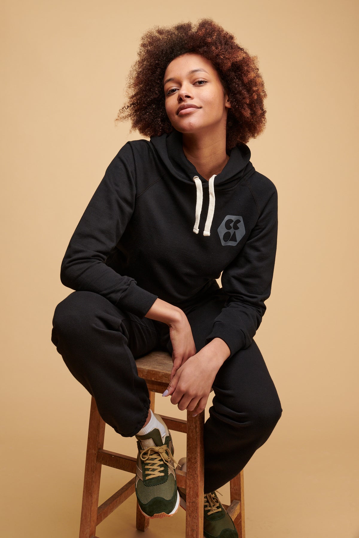 Female of colour sat on stool wearing plastic free hooded sweatshirt in black with ecru drawcord through the hood and CCOA logo print on chest paired with 100% cotton sweatpants in black