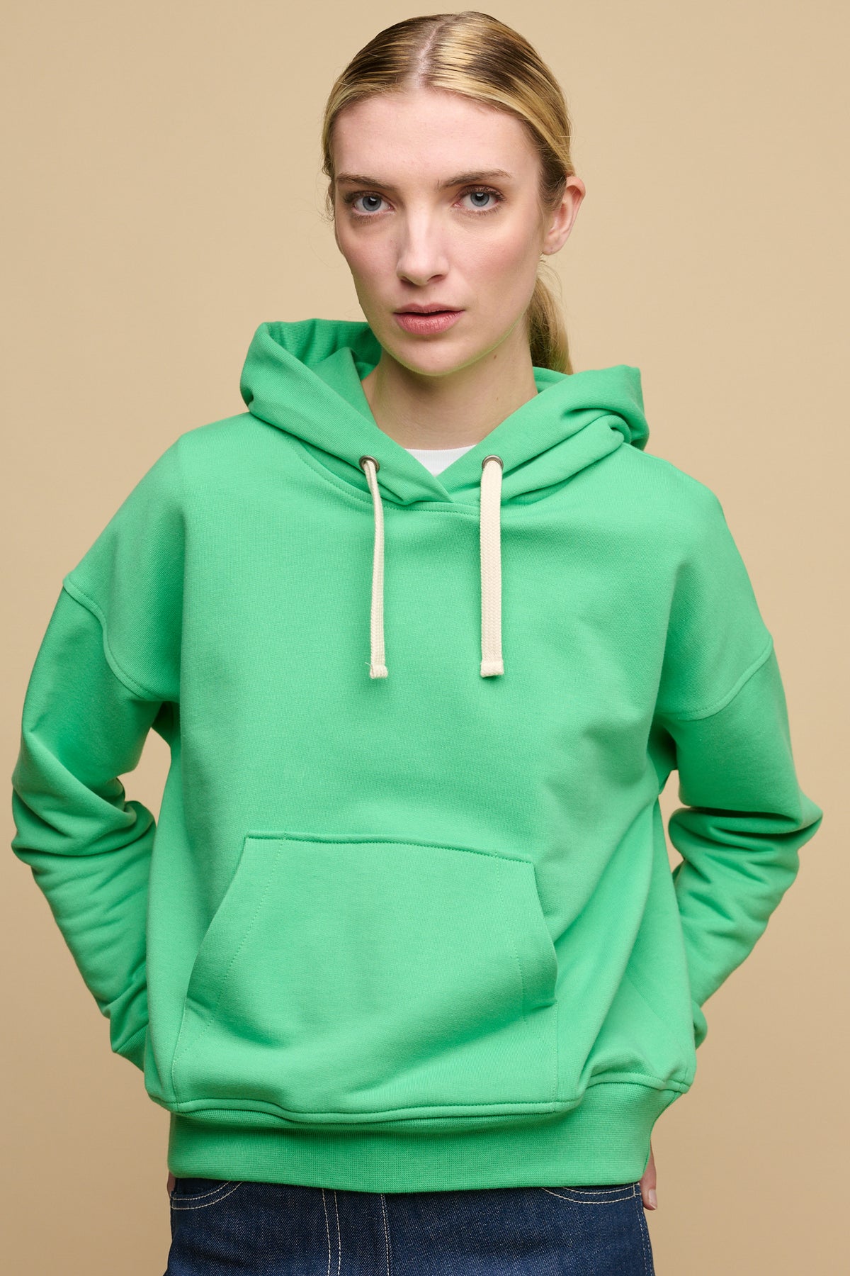 Thigh up image of female wearing hooded sweatshirt in apple green worn over white crew neck t shirt with blue jeans.